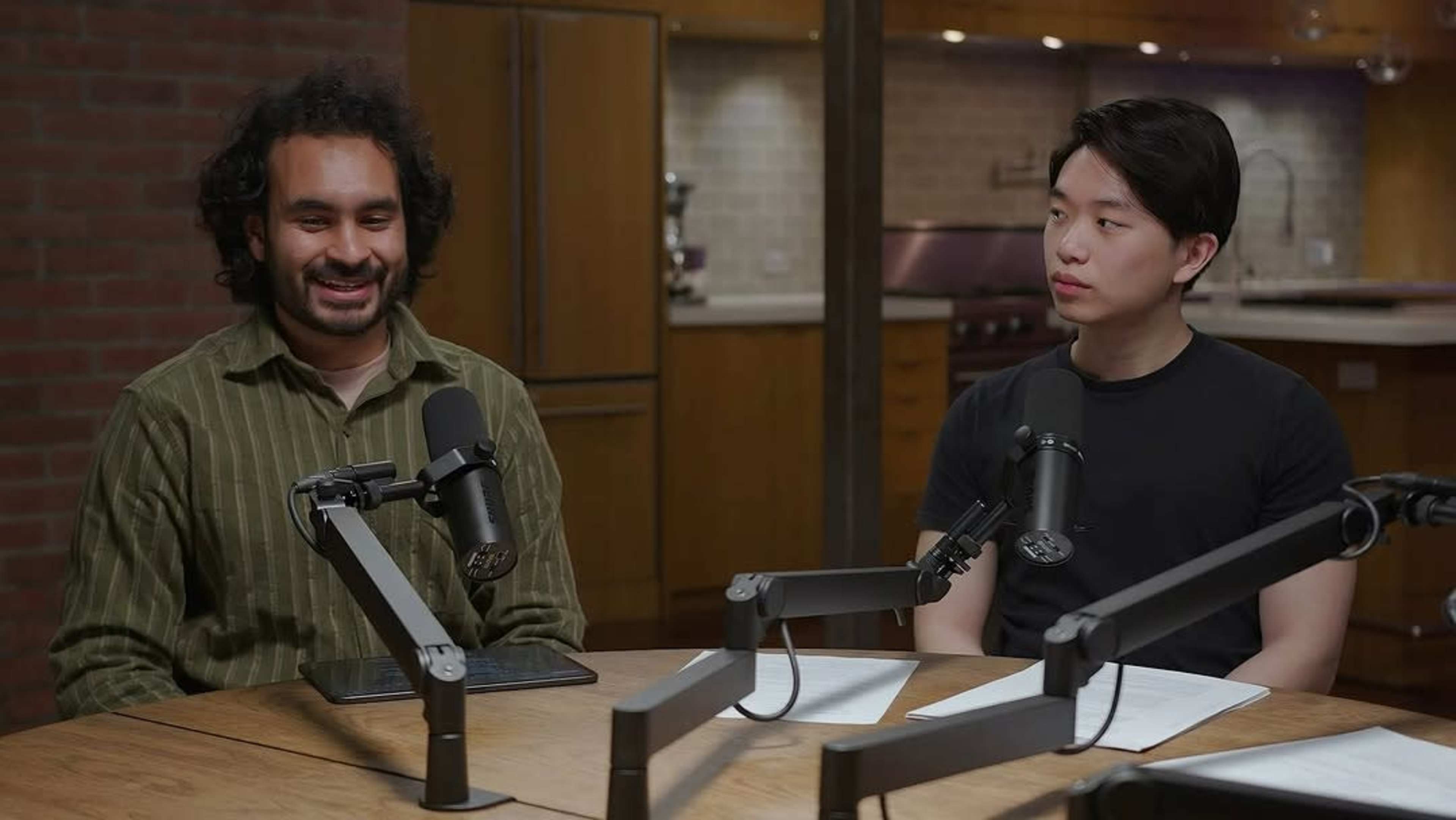 Two men are seated at a wooden table equipped with microphones, discussing in a well-lit kitchen setting.