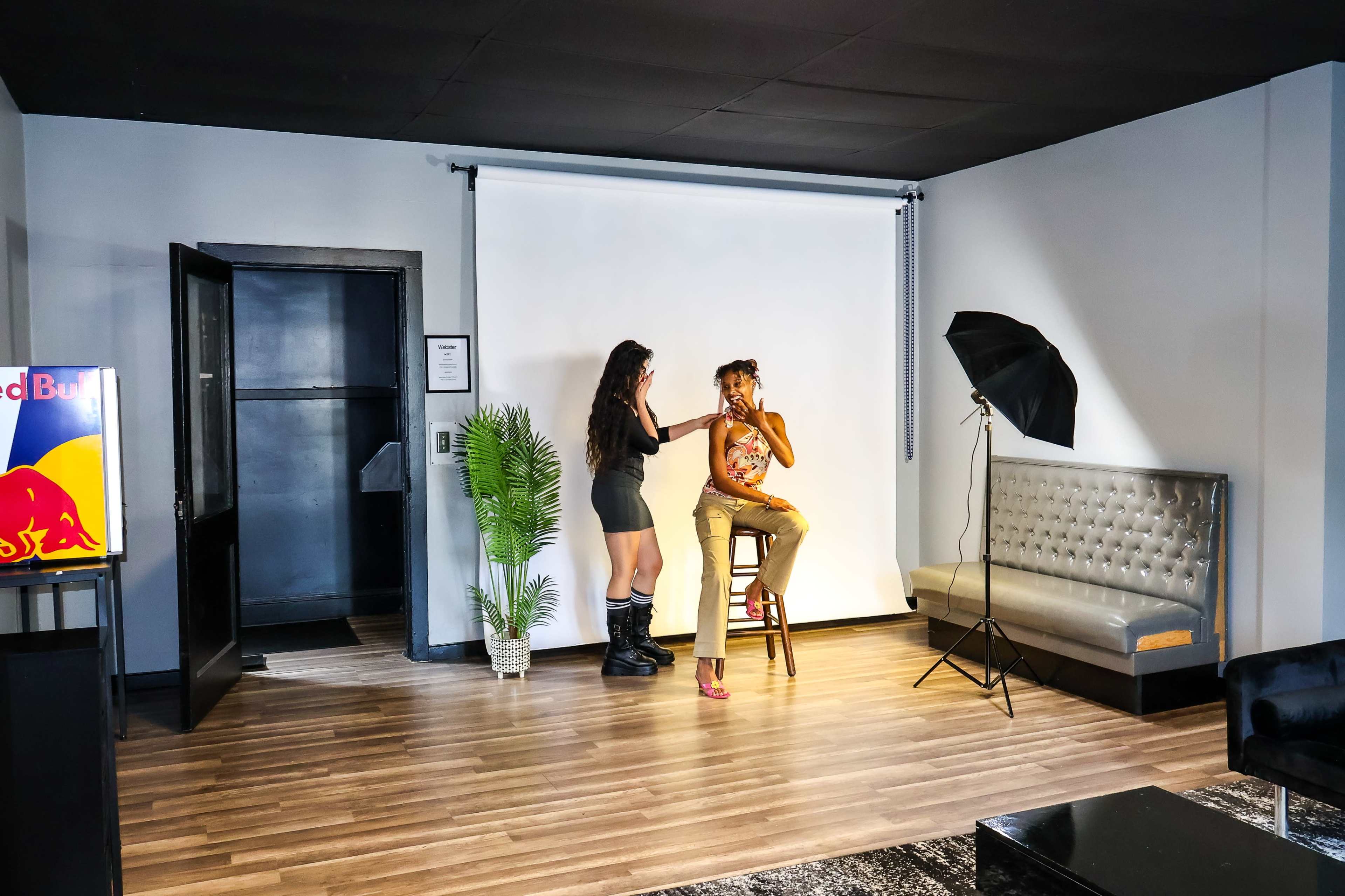 A woman in a black dress styles the hair of another woman sitting on a stool in front of a backdrop in a studio setting.