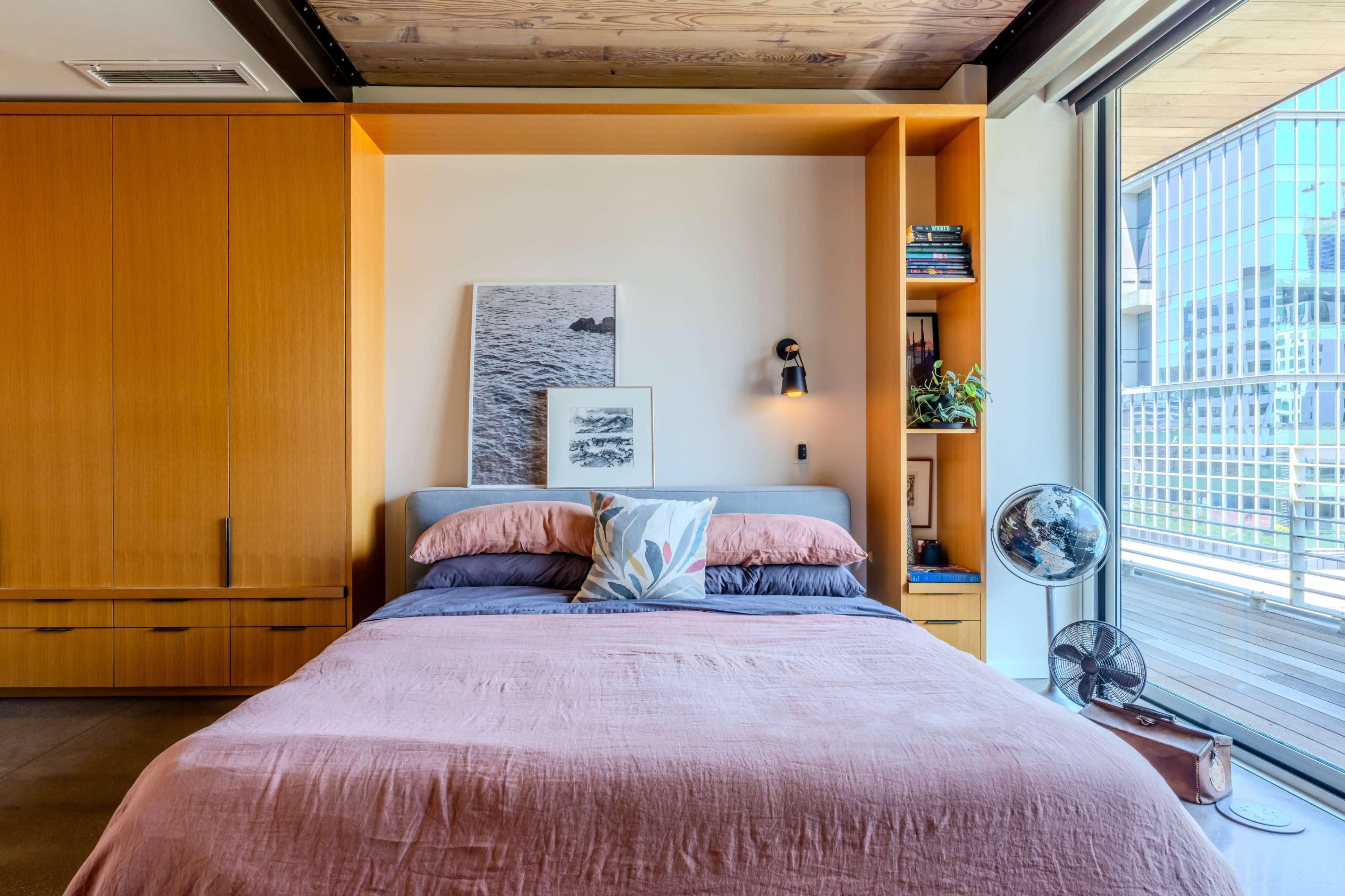 A neatly arranged bedroom features a bed with layered bedding, a wooden accent wall, and a shelf displaying books and decorative items.