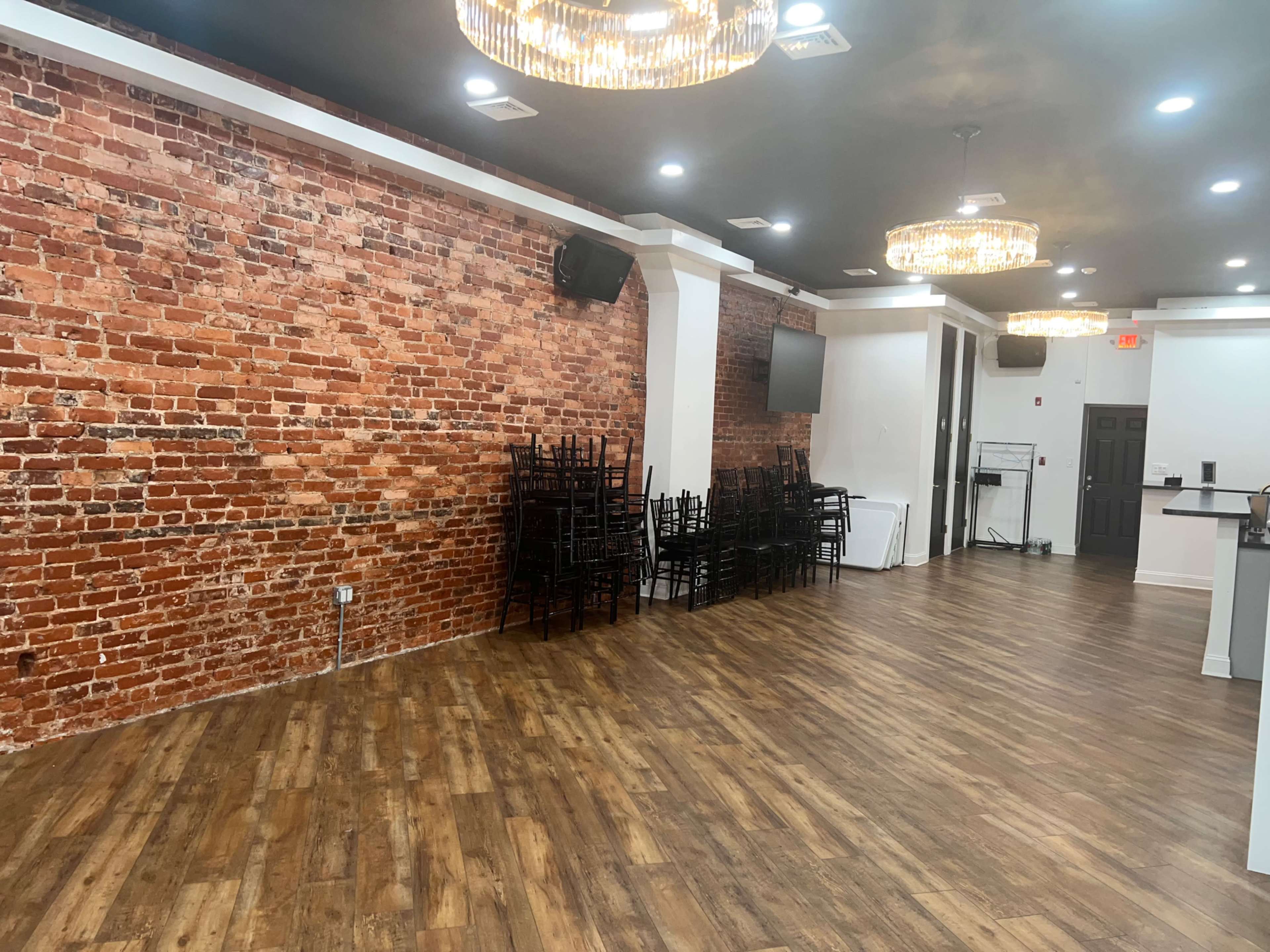 A spacious room with a brick wall, wooden flooring, and several stacked chairs under elegant chandeliers.