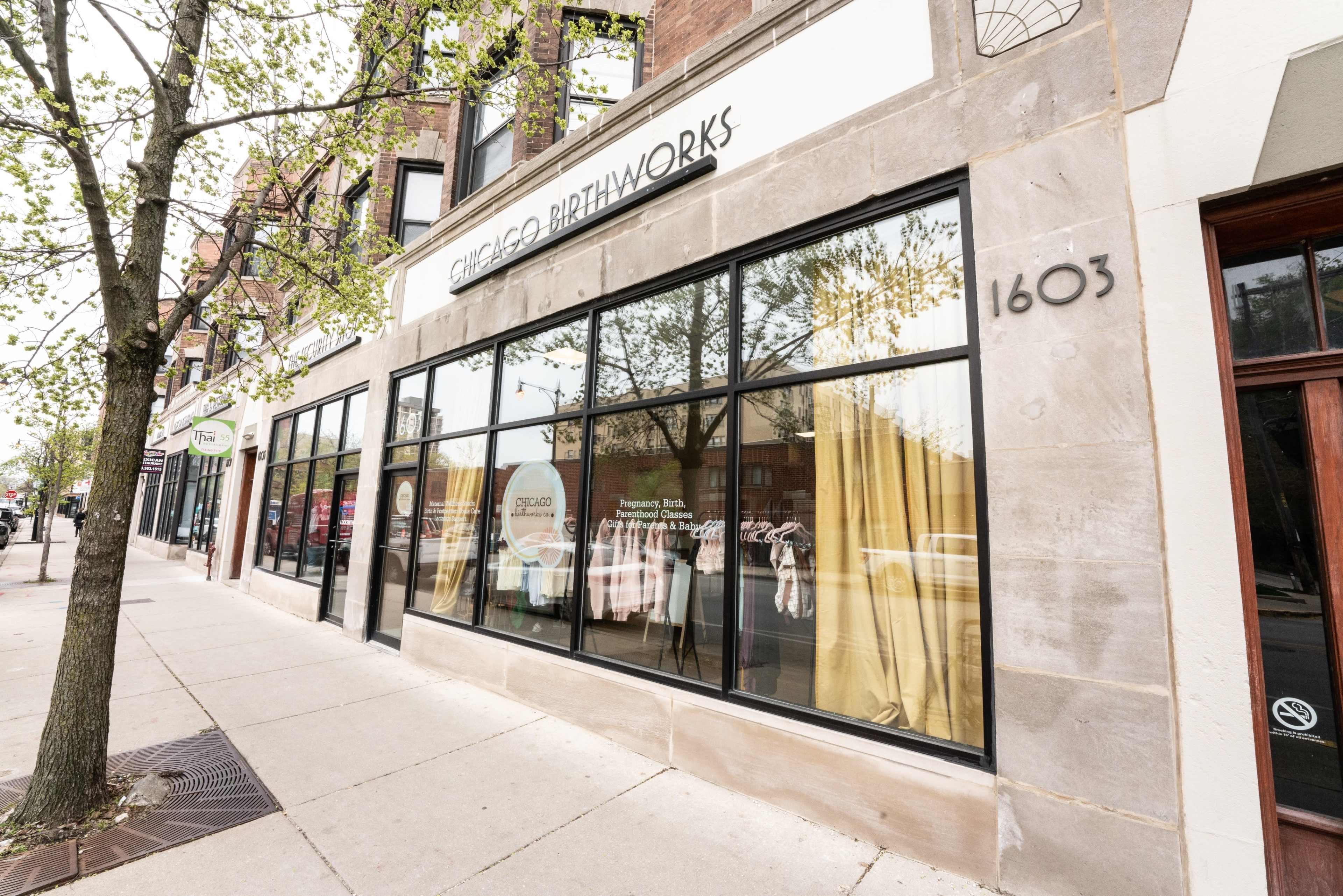 The storefront of Chicago Birthworks, featuring large windows with curtains and merchandise displayed inside, situated on a tree-lined sidewalk.