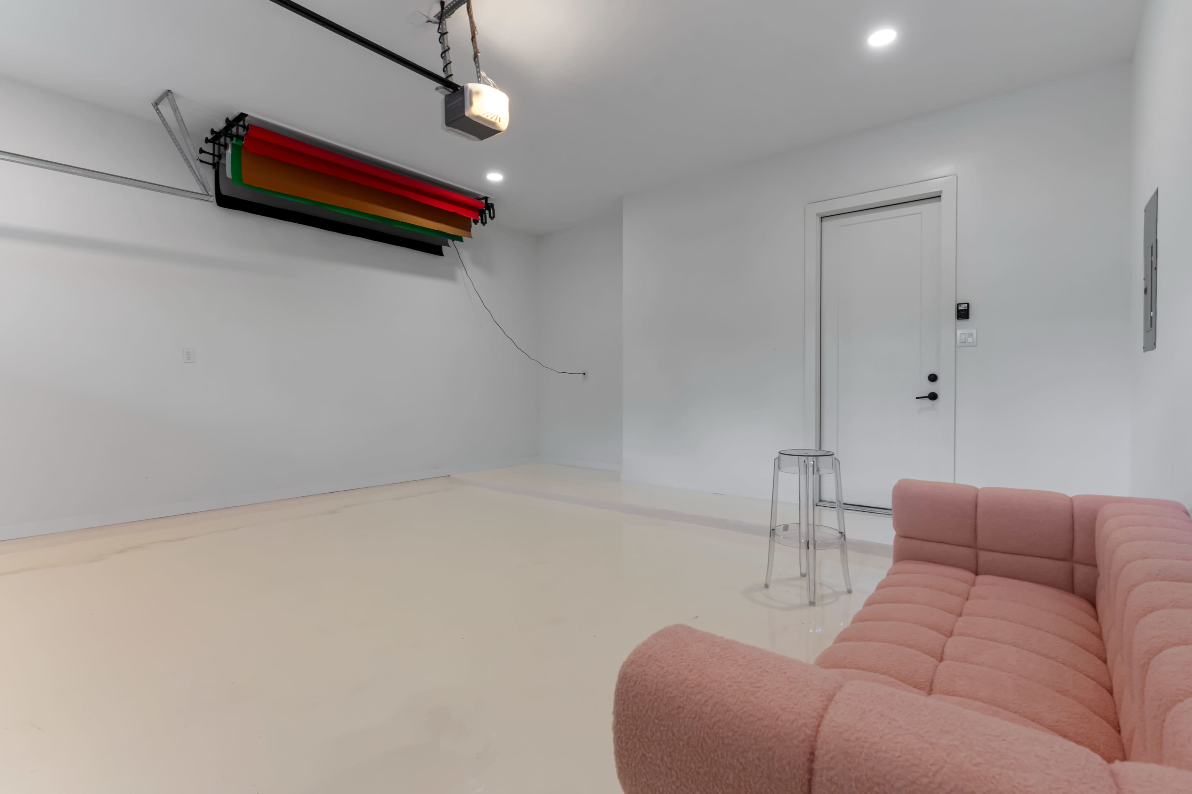The image depicts a minimalist interior space with a pink couch, a clear stool, and a door on one wall, while a retractable curtain system is mounted on the ceiling.