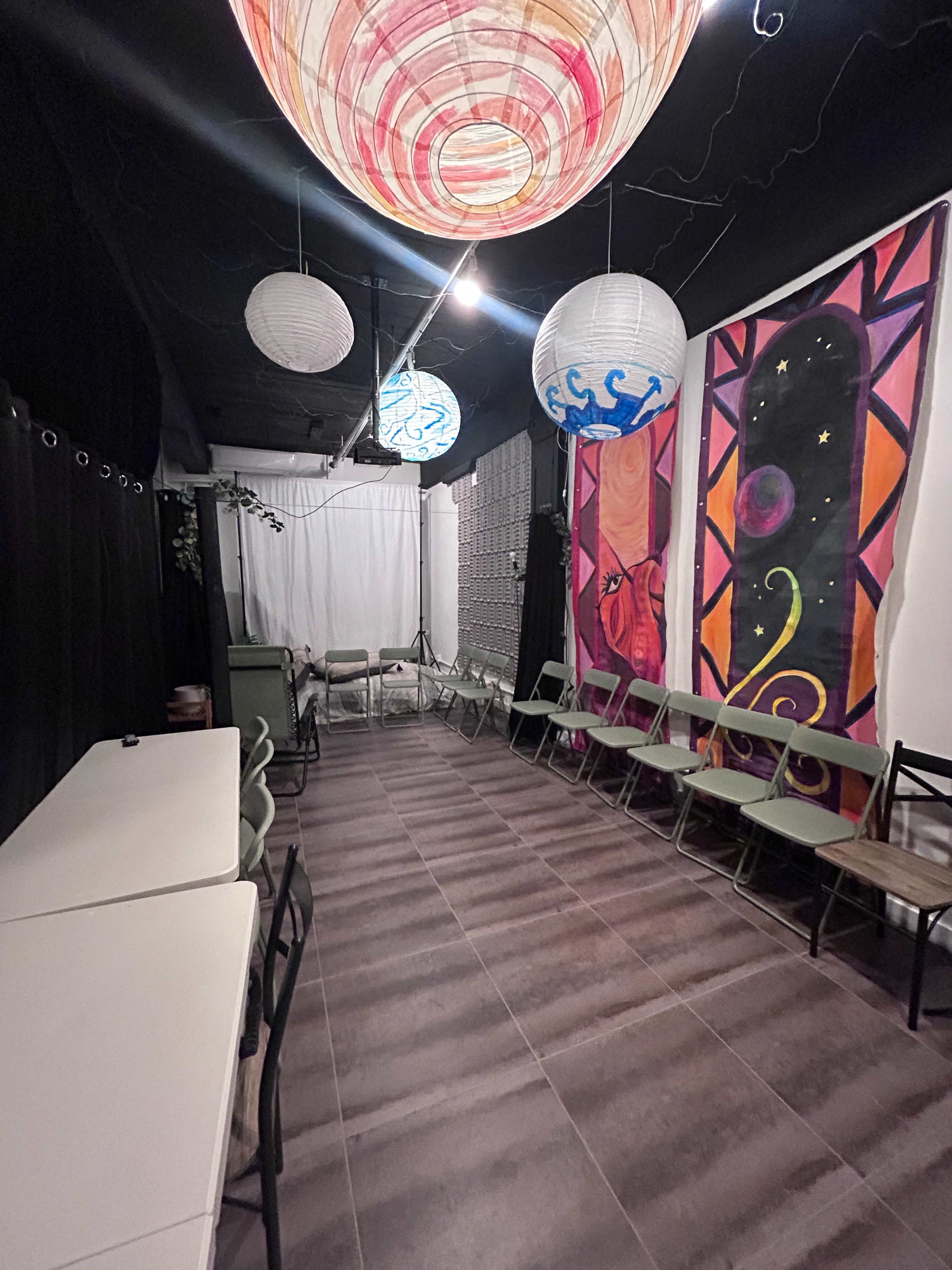 The image shows a long, narrow event space with folding chairs and tables, illuminated by colorful paper lanterns and featuring a brightly painted mural on one wall.