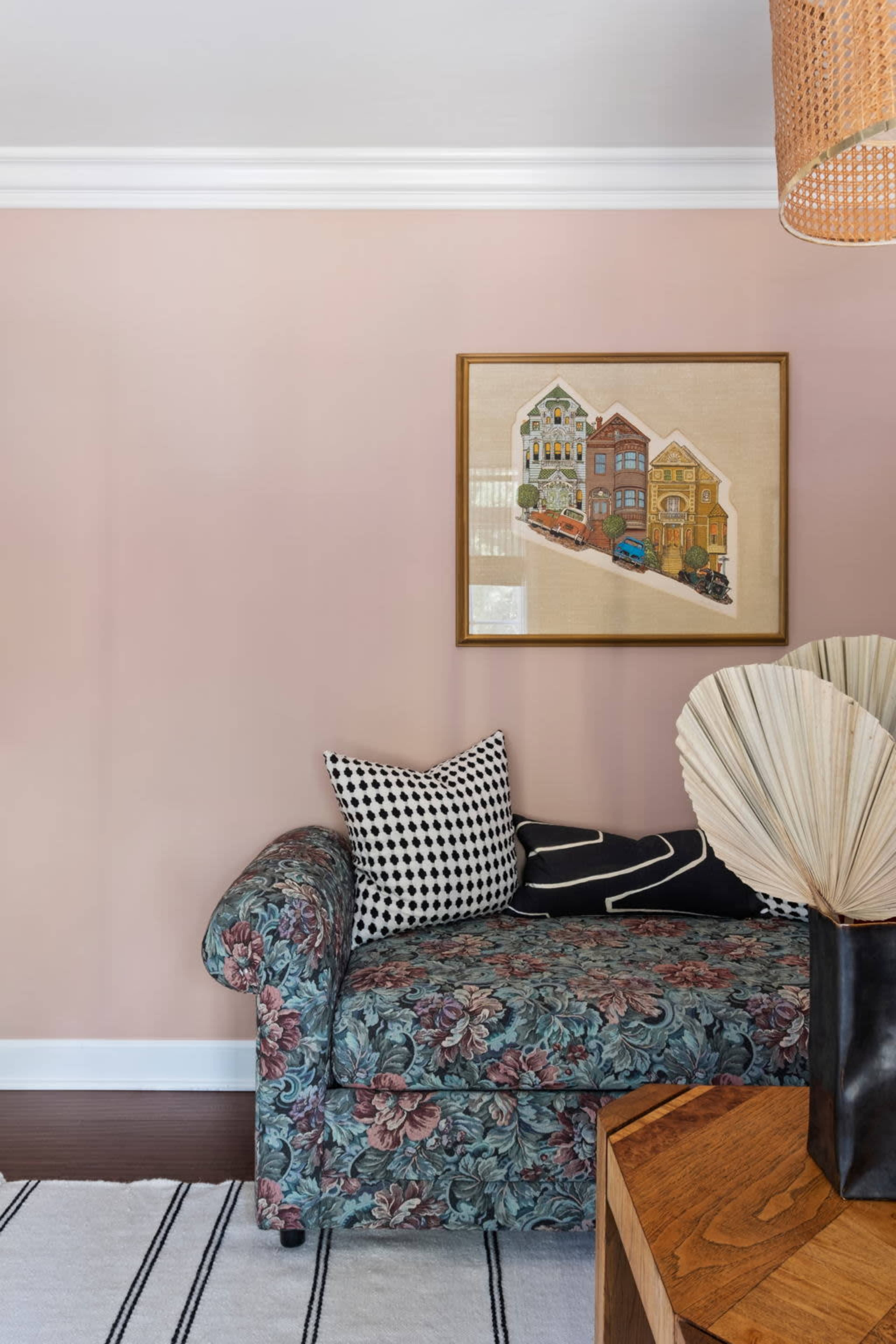 A floral patterned couch with black and white pillows is positioned against a pale pink wall, next to a wooden table and a decorative fan vase.