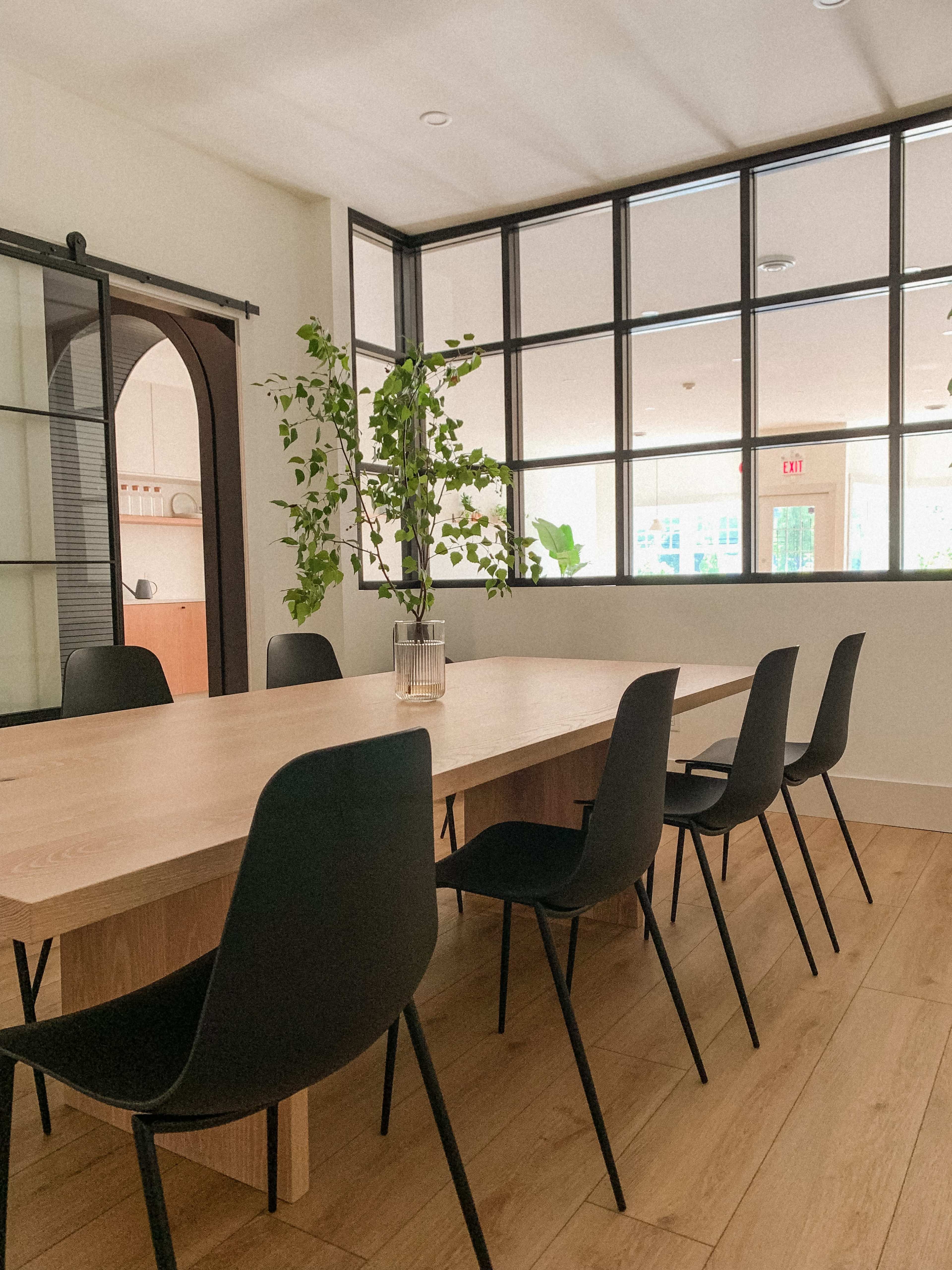 A large wooden table with black chairs surrounds it in a bright room with large windows and potted plants.