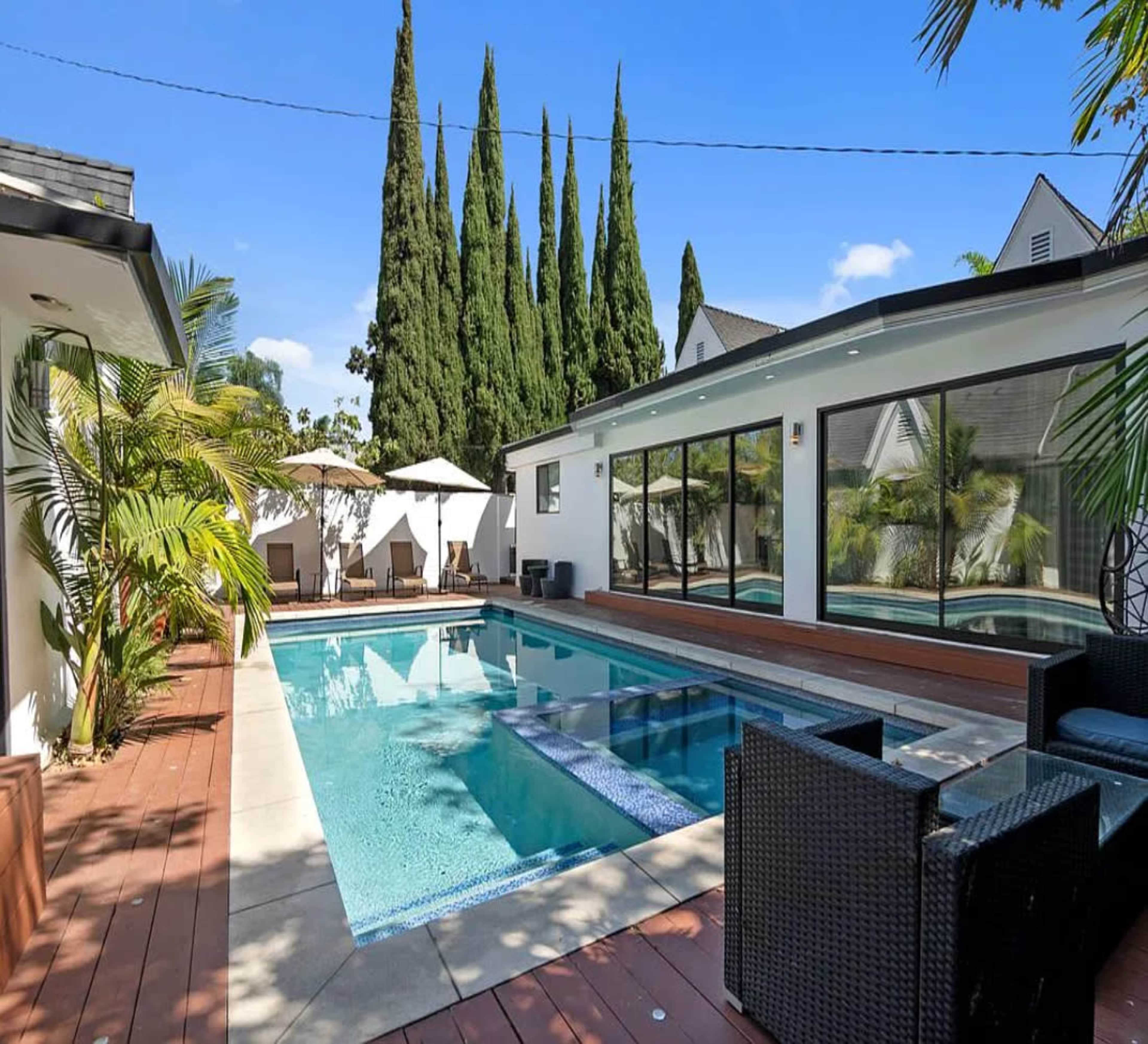 The image shows a modern swimming pool surrounded by a wooden deck, lounge chairs, and tall cypress trees under a clear blue sky.