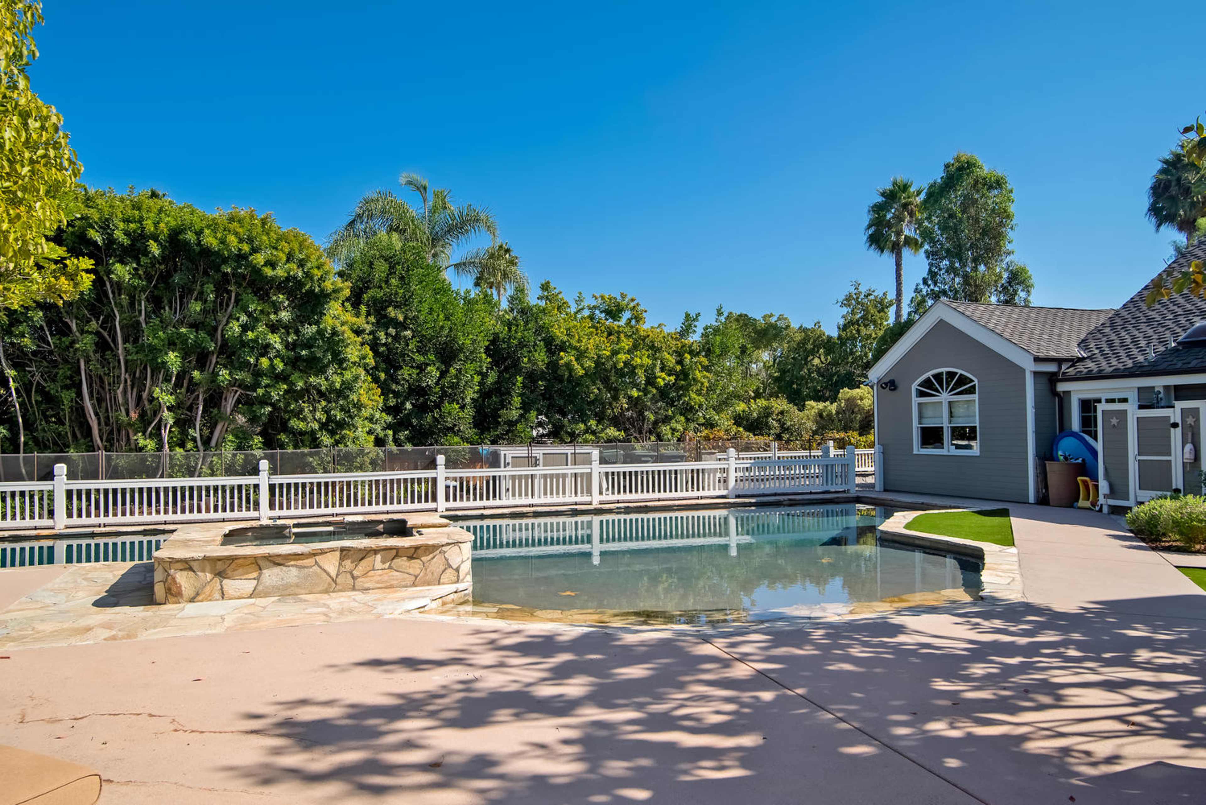 The image shows a landscaped backyard with a swimming pool, a stone hot tub, and a gazebo surrounded by lush greenery.