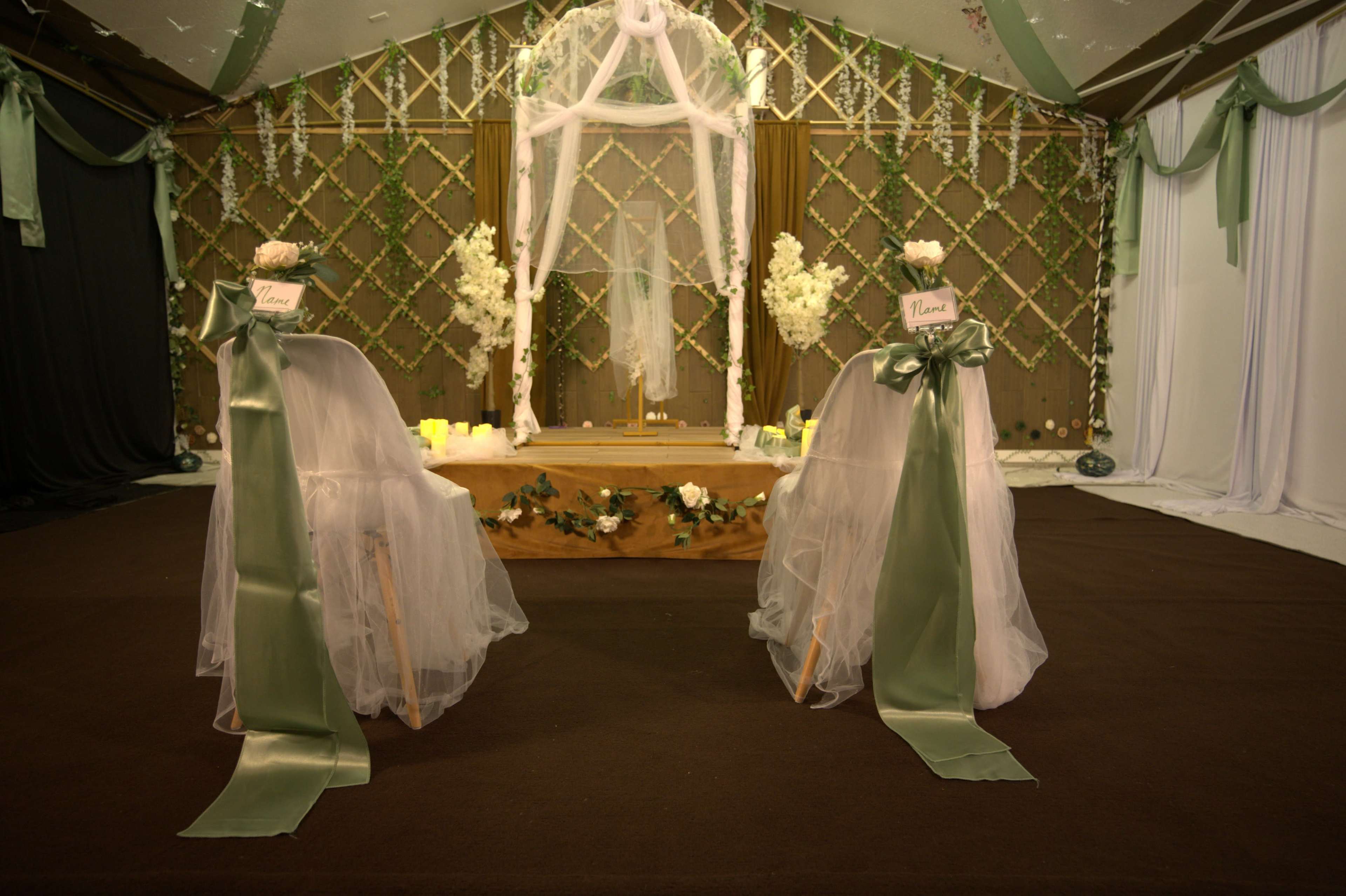 The image shows an elegantly decorated wedding altar with two chairs, each adorned with white tulle and green ribbons, set against a backdrop of greenery and soft lighting.
