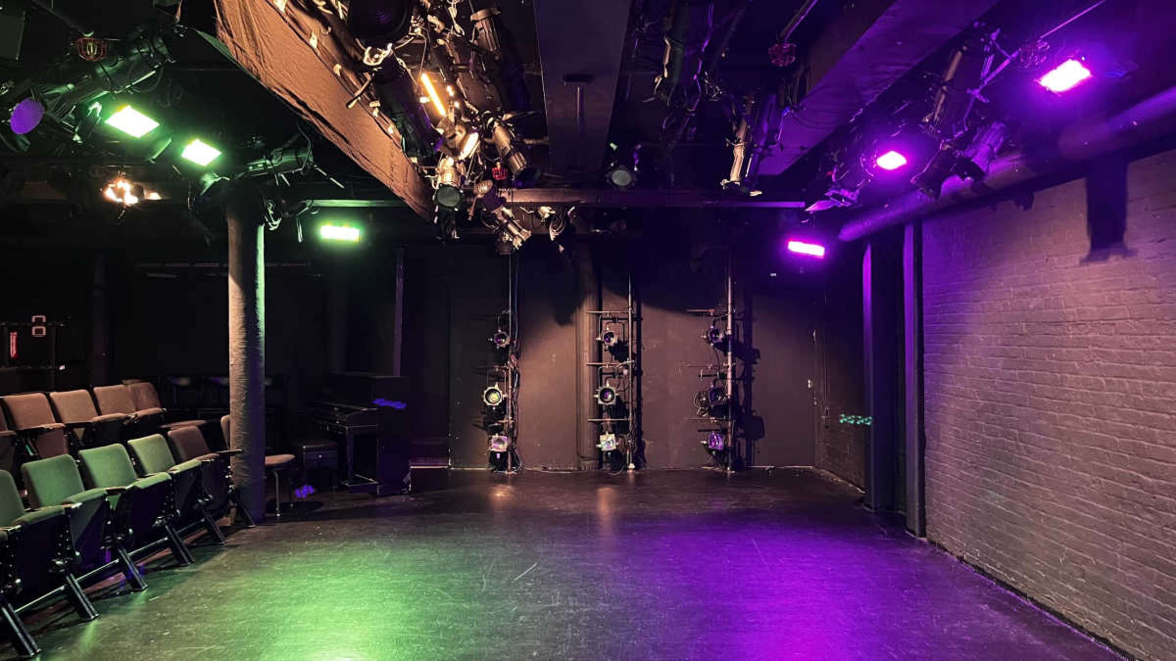 The image shows a dimly lit theater stage with seating on one side and various brightly colored stage lights mounted above.