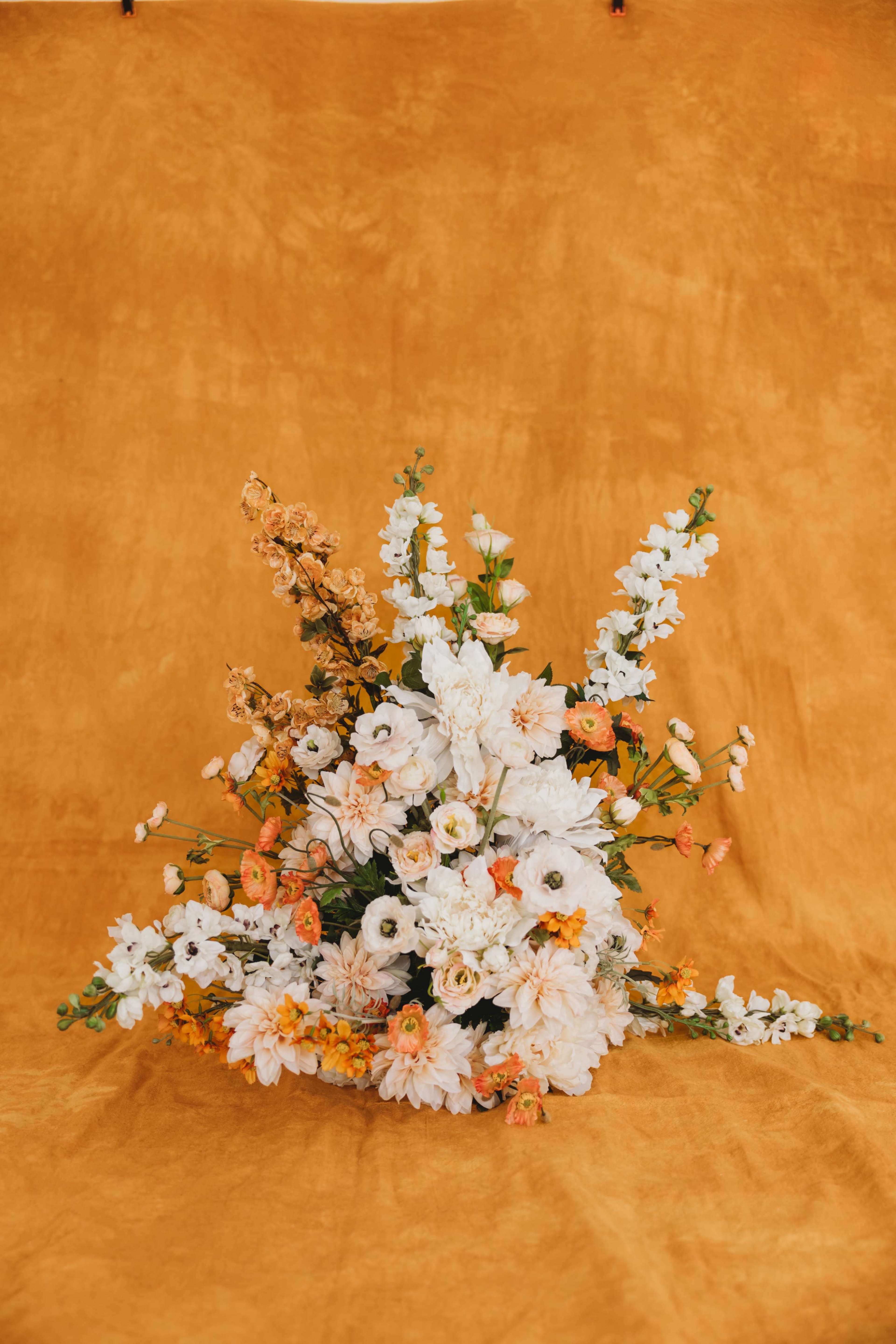 A floral arrangement featuring a variety of white and orange flowers is positioned in front of an orange backdrop.