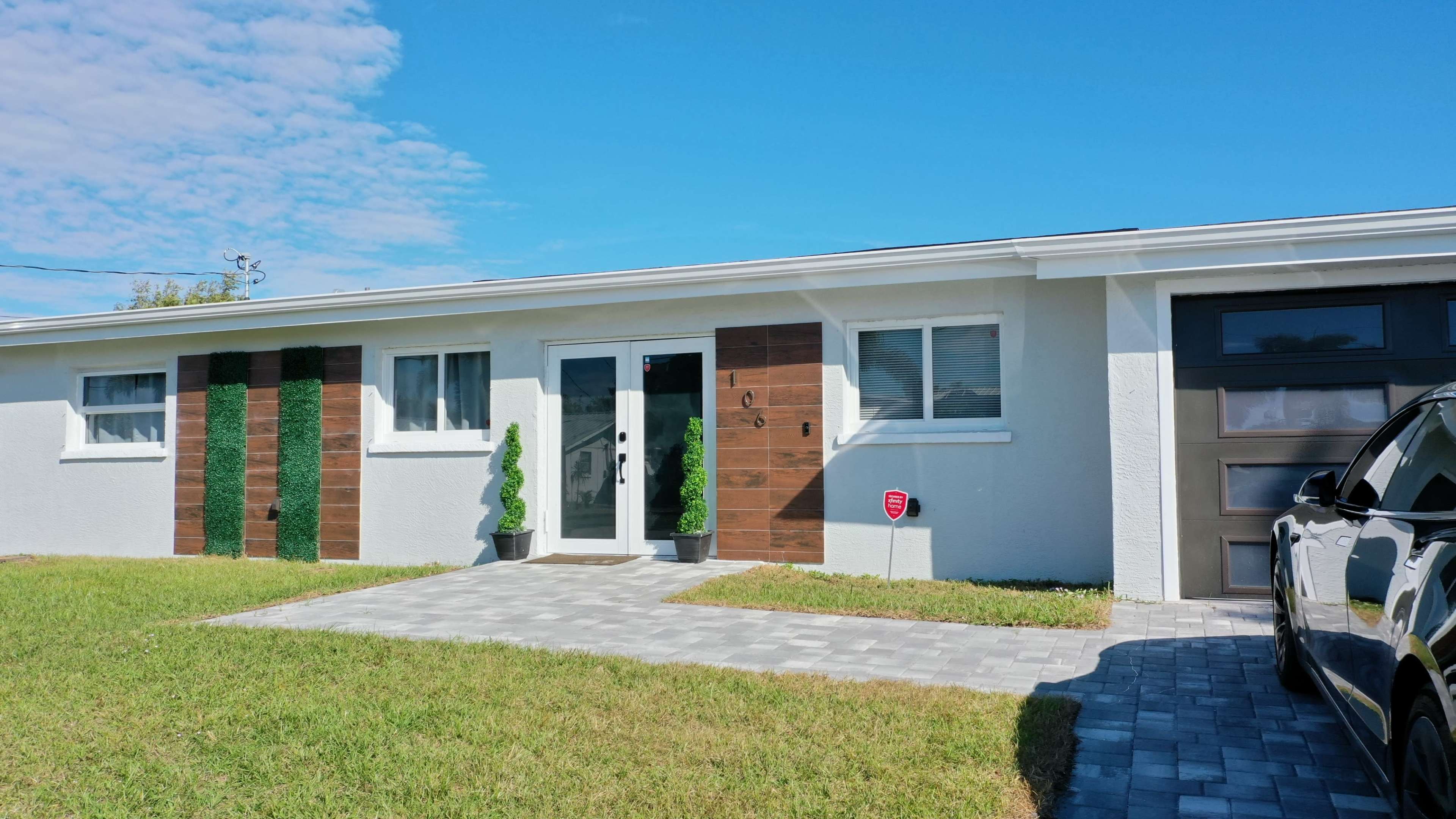 The image shows a modern house with a light-colored exterior, a wooden accent wall, and landscaped grass in the front yard.