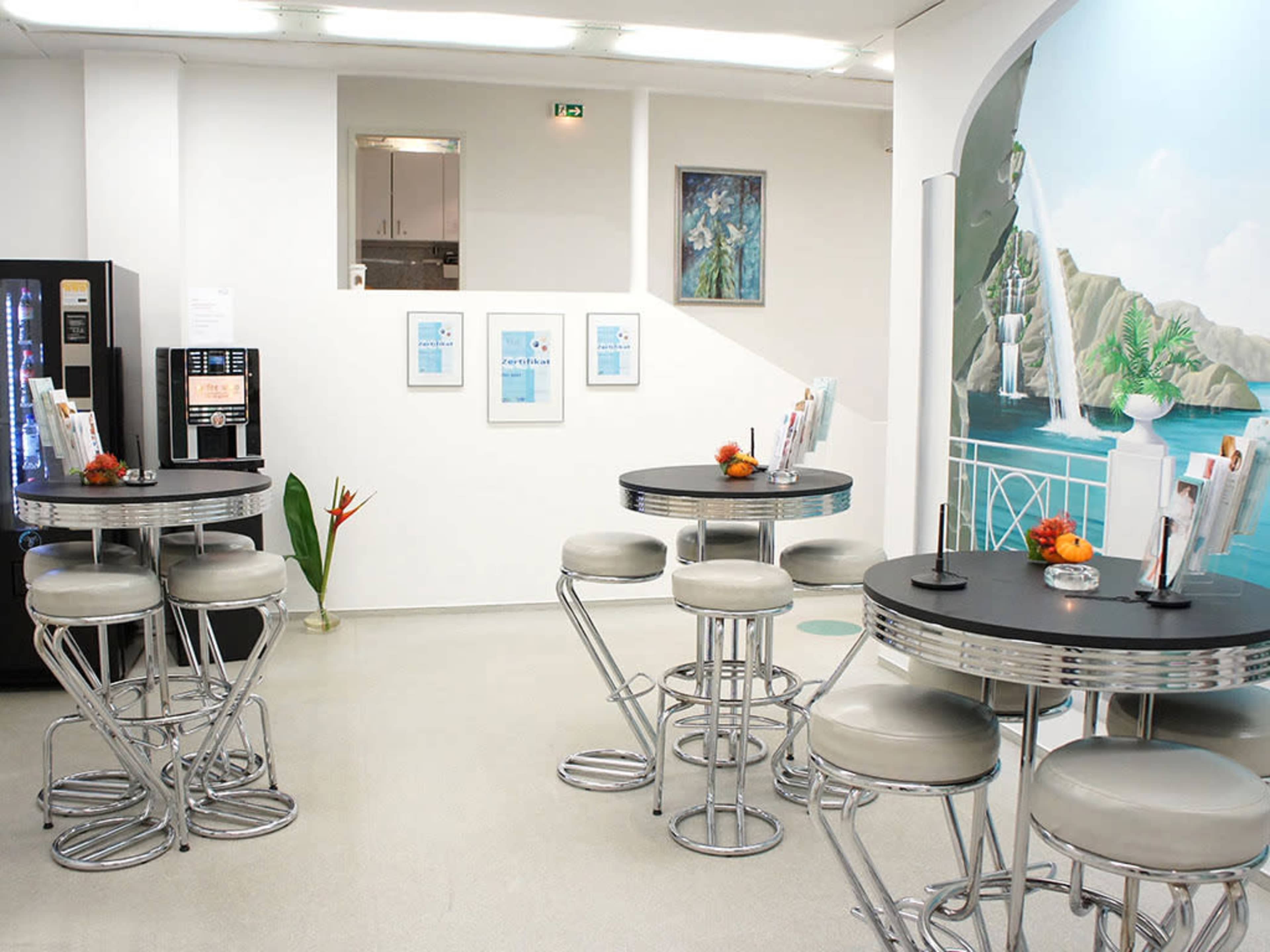A modern, bright indoor space featuring several high round tables with metal and upholstered stools, surrounded by minimalistic decor, including a vending machine and wall art.