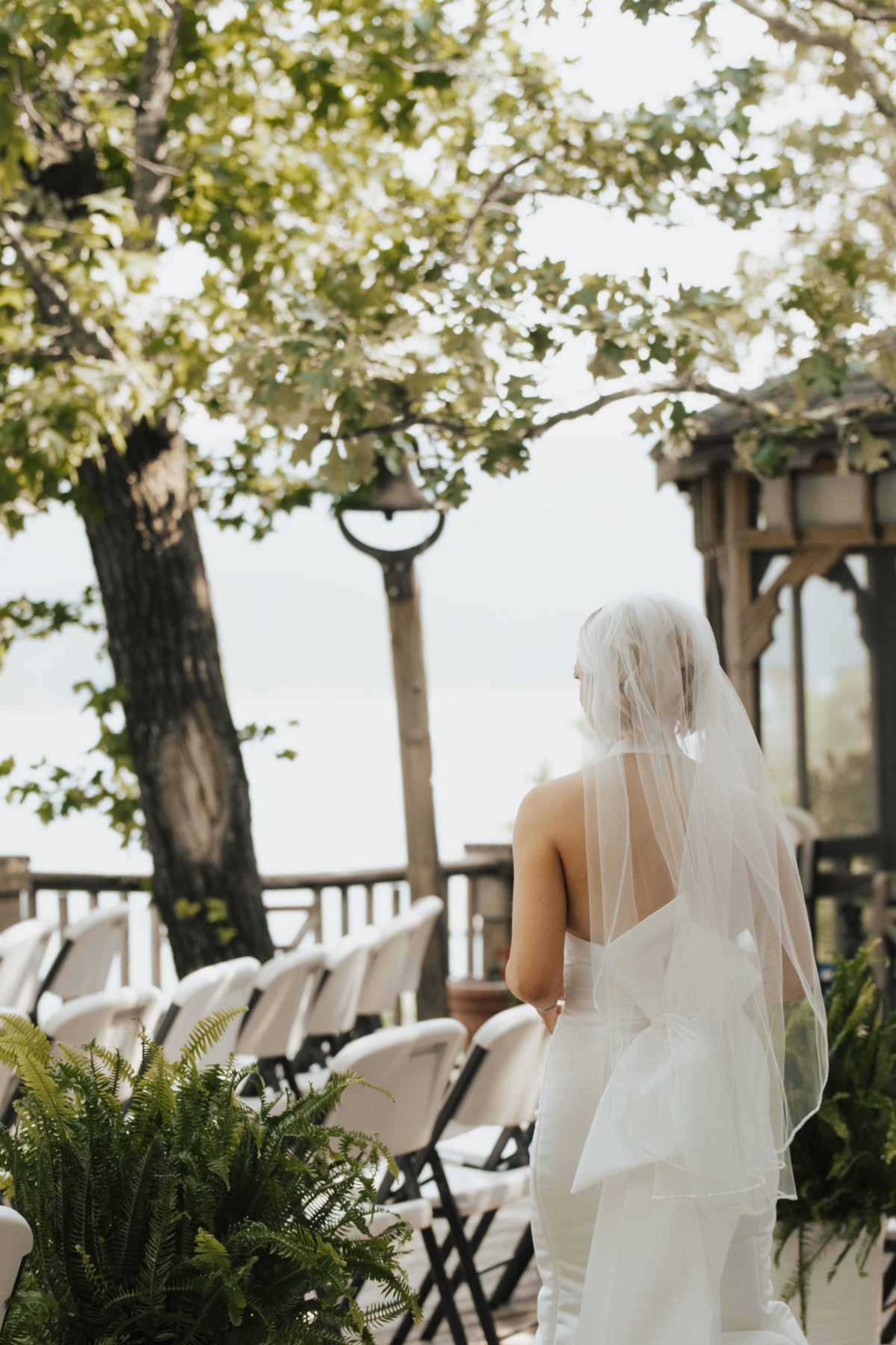 A bride in a backless white dress stands with her back to the camera, surrounded by chairs arranged for a wedding ceremony under leafy trees.