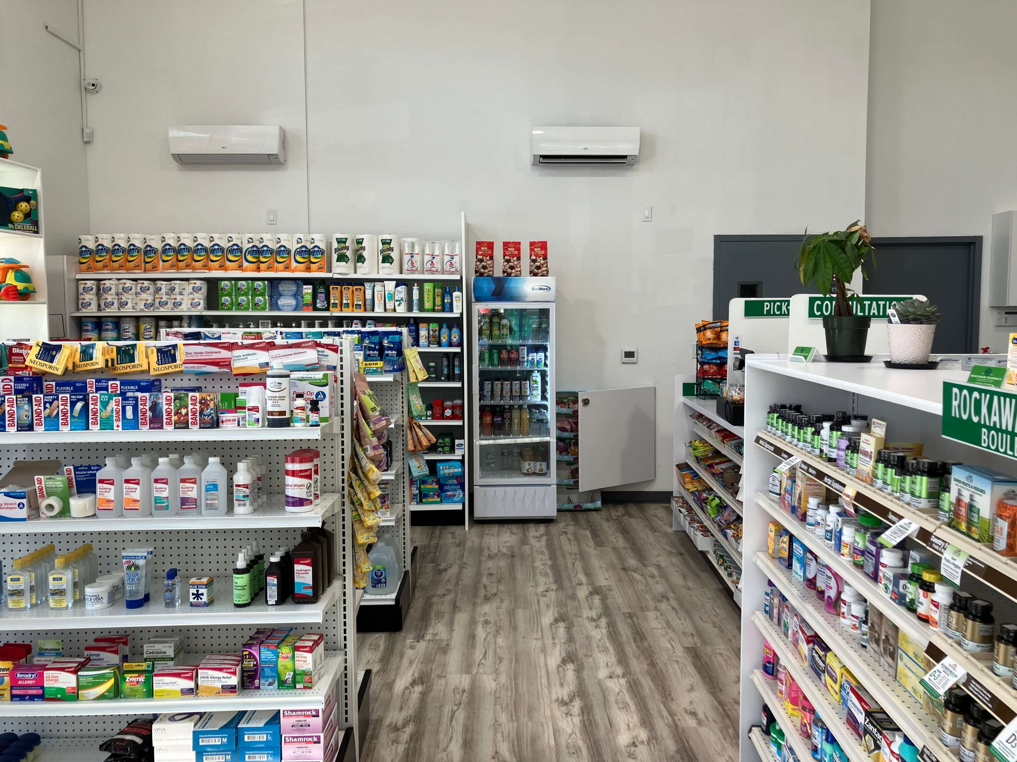 The image shows the interior of a pharmacy with shelves stocked with various medications and health products, along with a refrigerated section in the center.