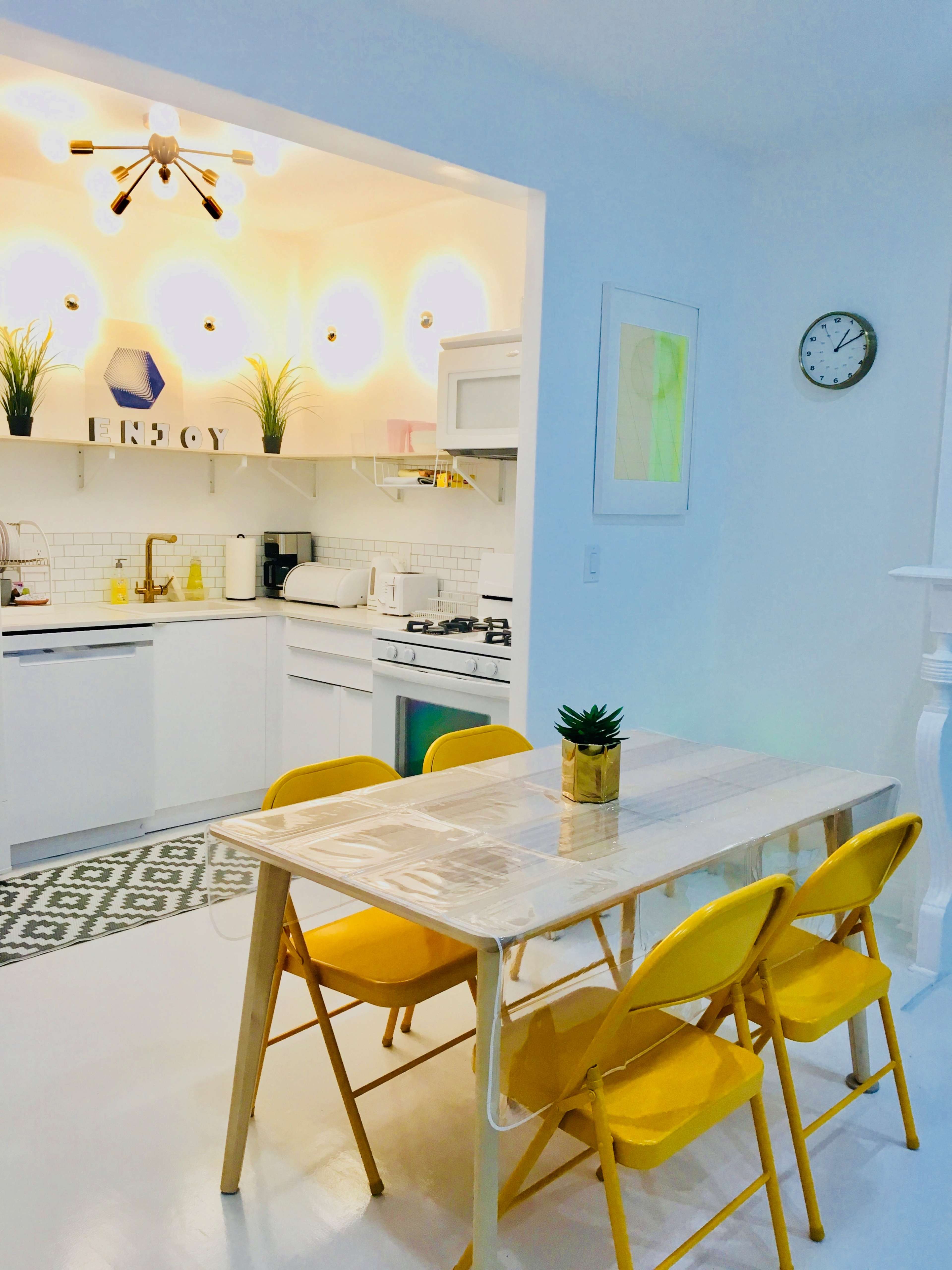 A bright, modern kitchen features a dining table with yellow chairs and a view of white cabinetry, plants, and a wall clock.