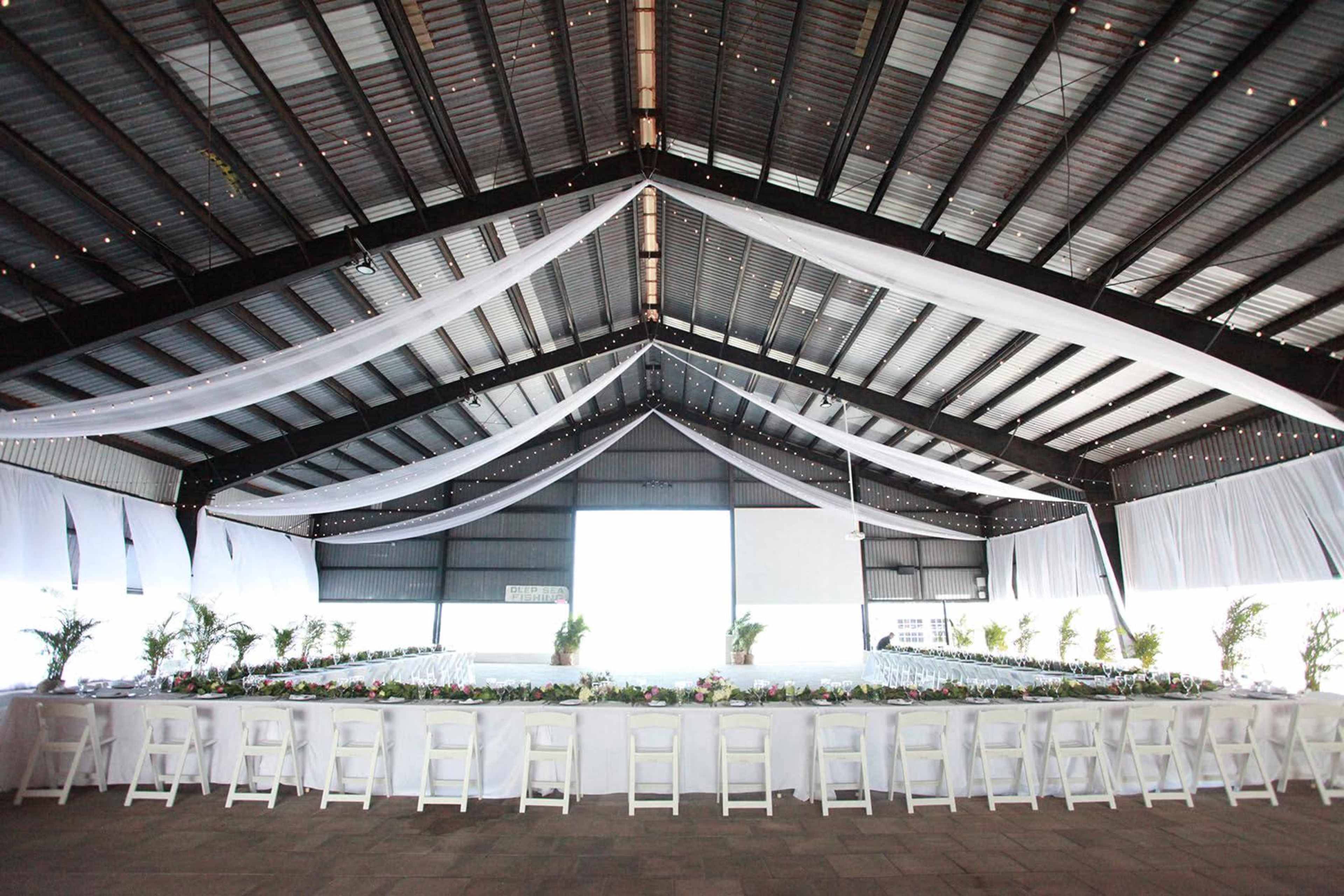 A large indoor venue with a long table set for an event, featuring white tablecloths and floral arrangements along the center, beneath a high, draped ceiling with string lights.