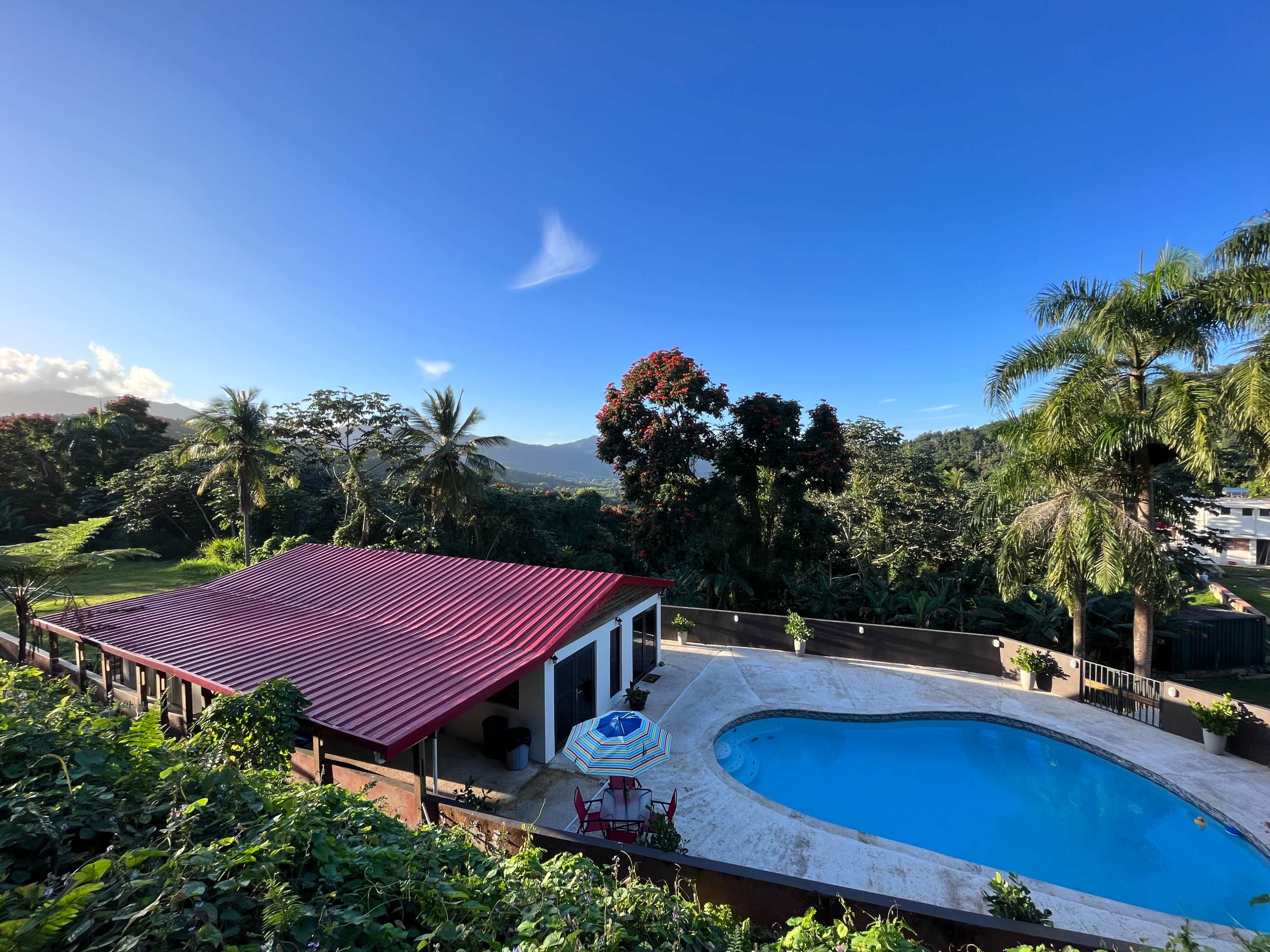 The image shows a house with a red roof beside a swimming pool, surrounded by tropical greenery and mountains in the background.
