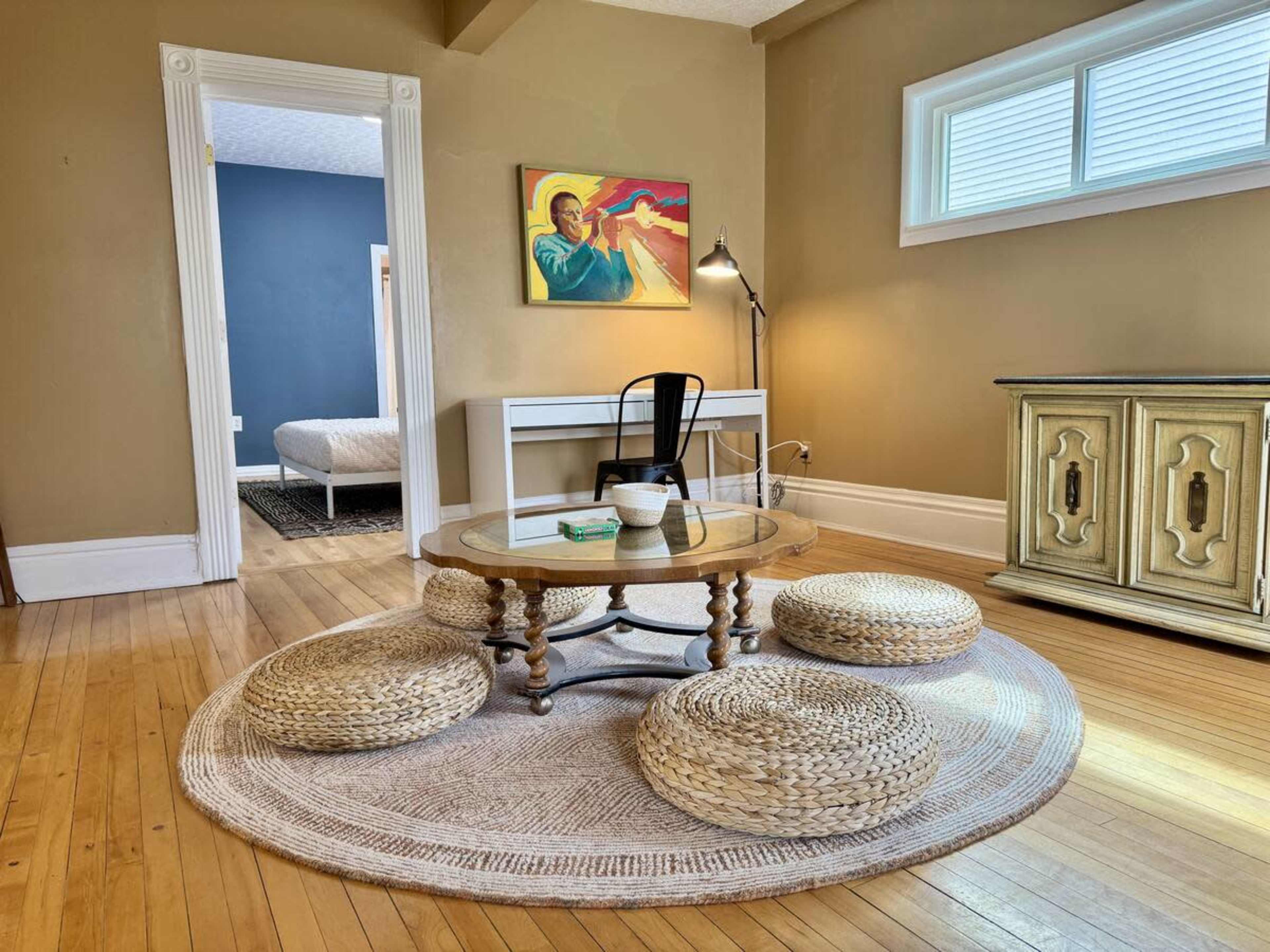 The image shows a living space with a round woven rug, a wooden coffee table, and several floor cushions, alongside a desk and a painting on the wall.