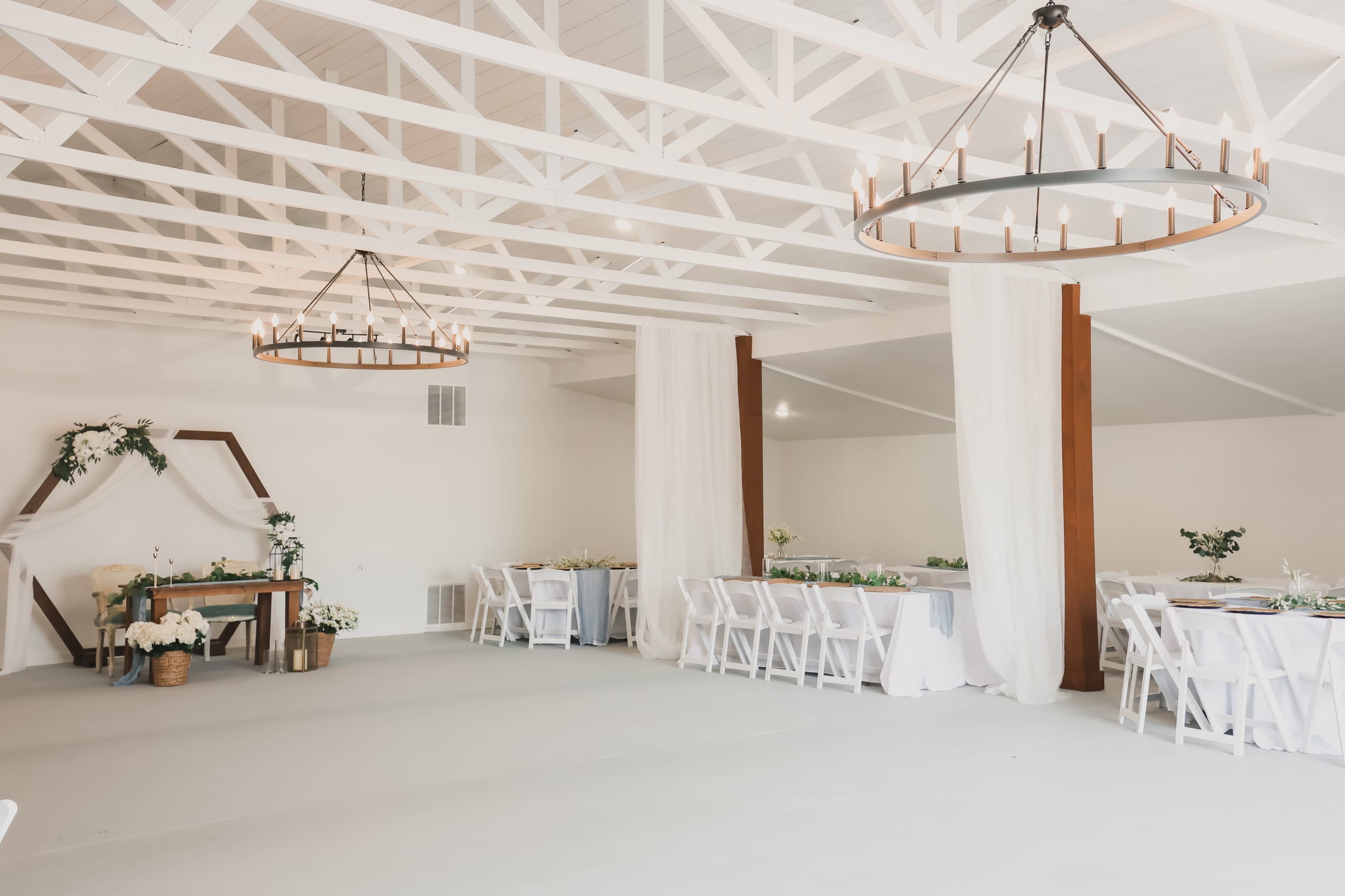 The image shows a spacious event venue featuring white walls, exposed beams, and two chandeliers, with tables set for a gathering and a decorative arch in one corner.