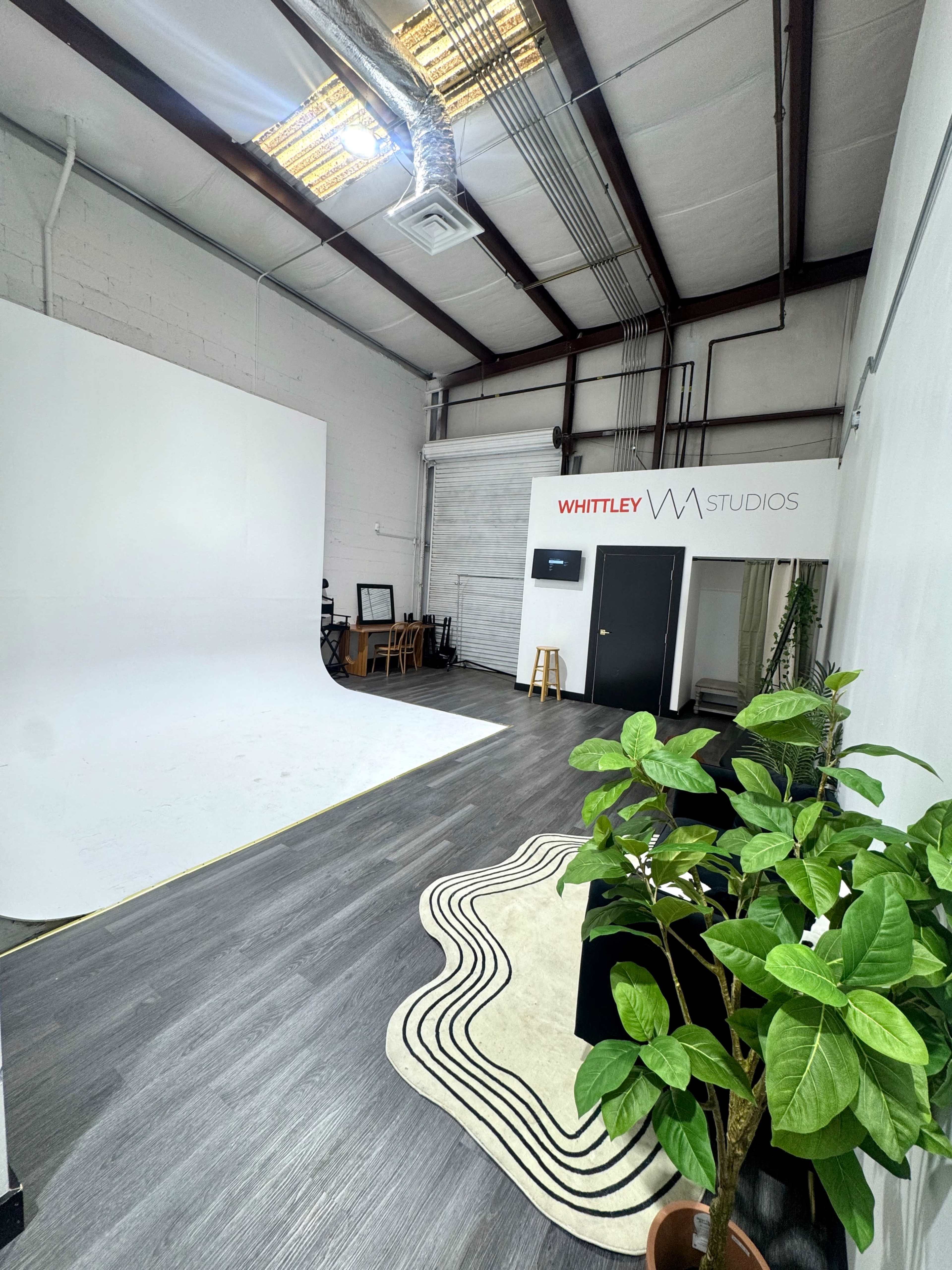 The image shows a spacious photography studio featuring a large white backdrop, a seating area with wooden furniture, and a decorative plant beside a patterned rug.