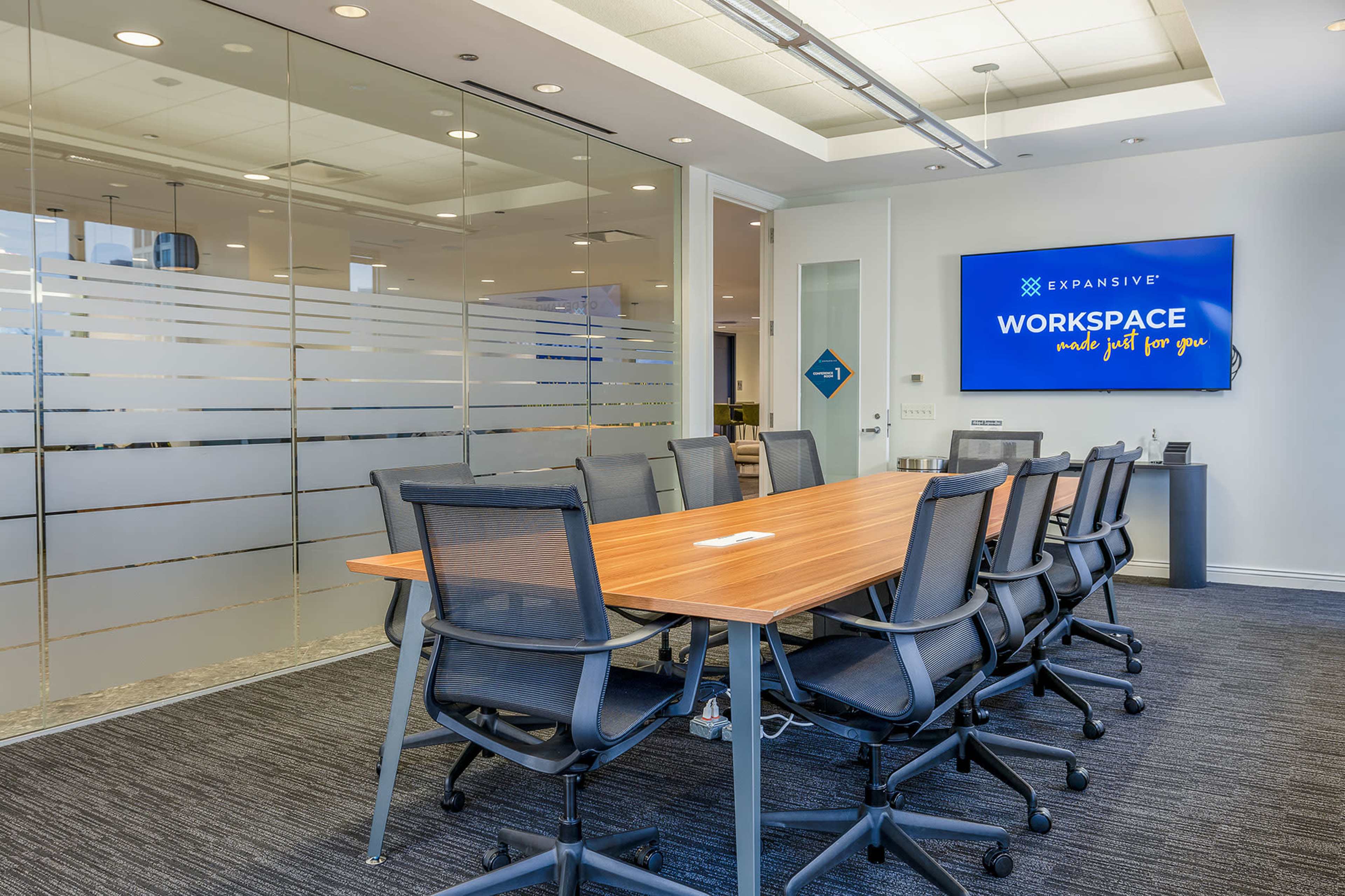 A modern conference room features a long wooden table surrounded by chairs, with a large screen displaying the word "WORKSPACE" and a glass wall in the background.