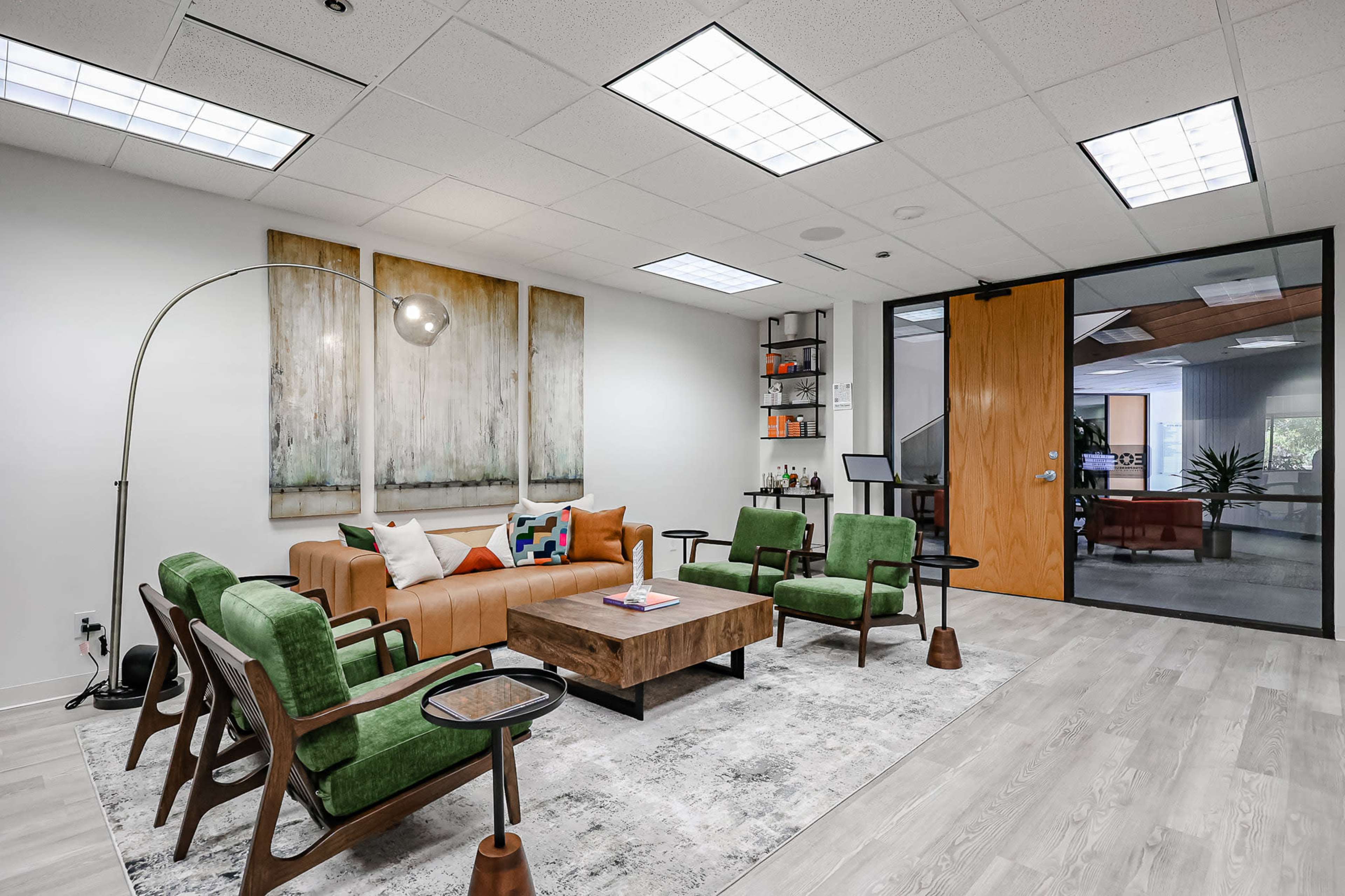 The image shows a modern office lounge area with a brown leather sofa, green armchairs, a wooden coffee table, and decorative wall art.