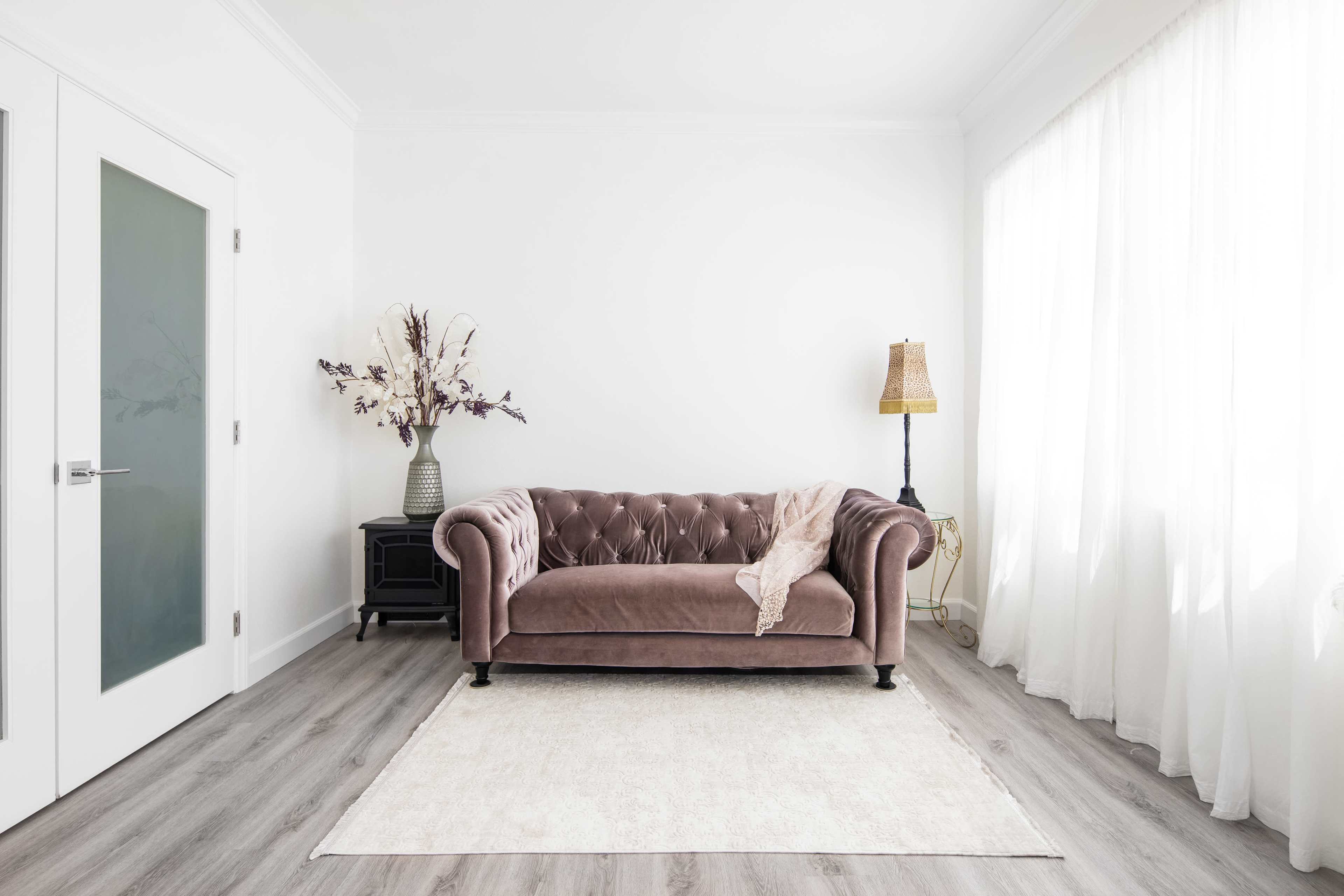 A tufted brown sofa sits in a minimalist living room with a white rug, a lamp, and a vase of dried flowers beside a glass door and sheer curtains.