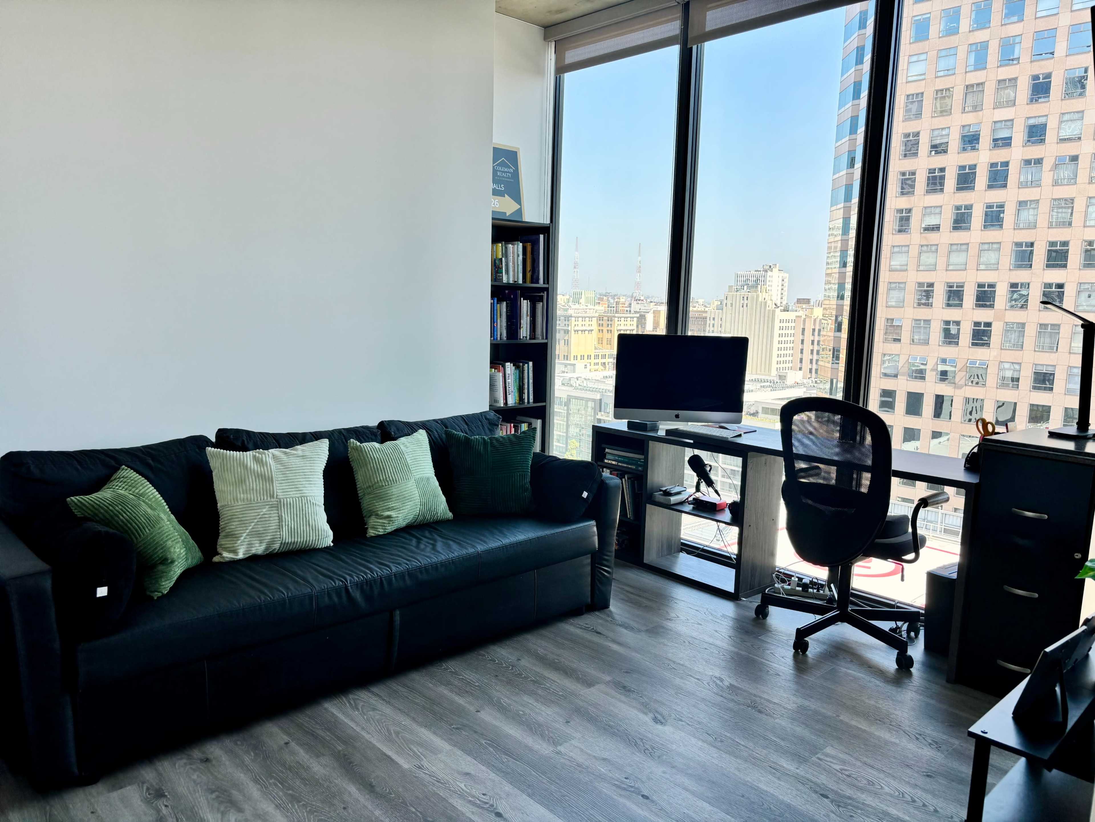 The image shows a modern office space featuring a black couch, a bookshelf, a desk with a chair, and large windows that provide a city view.