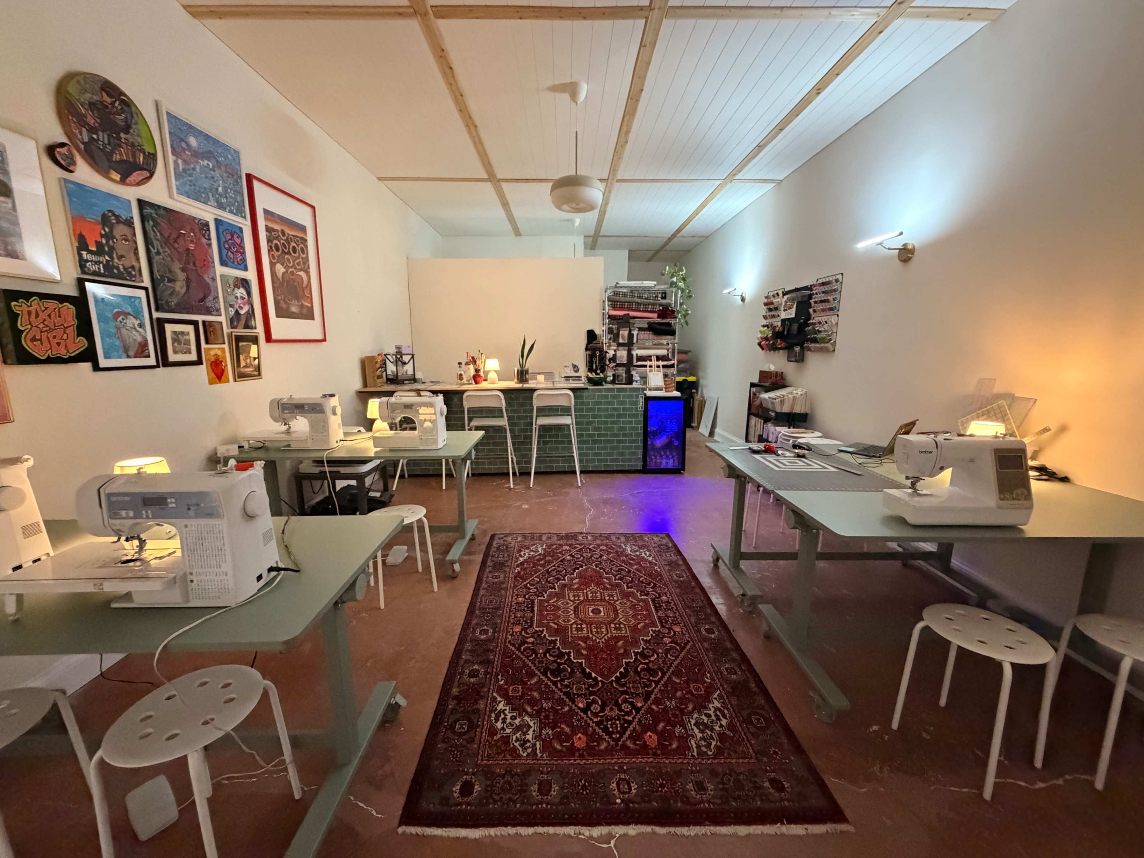 The image shows a bright sewing studio with multiple sewing machines arranged on tables, a patterned rug on the floor, and a counter with lamps and creative supplies in the background.