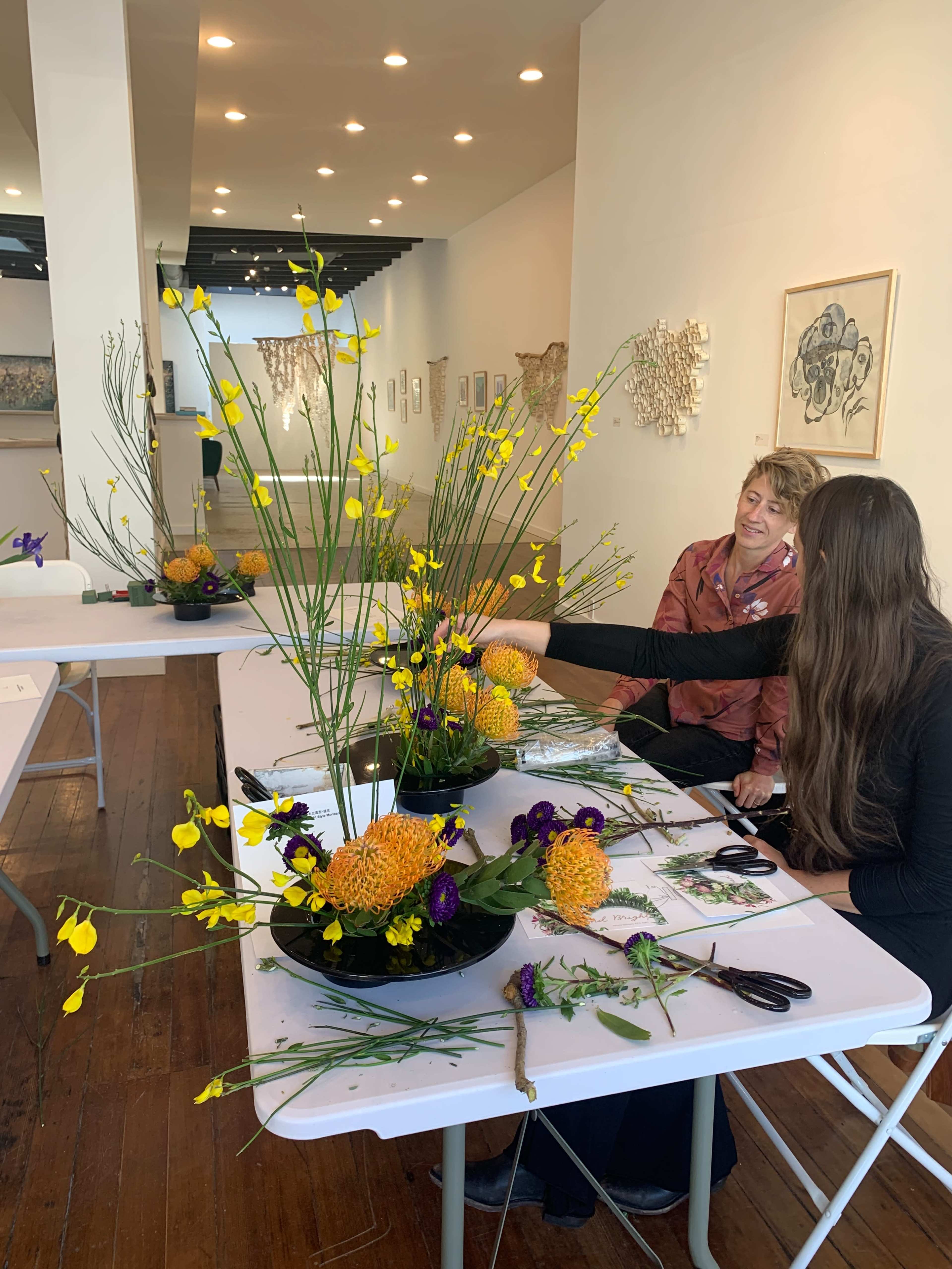 Two individuals are arranging floral displays on tables in an art gallery, surrounded by various artworks on the walls.