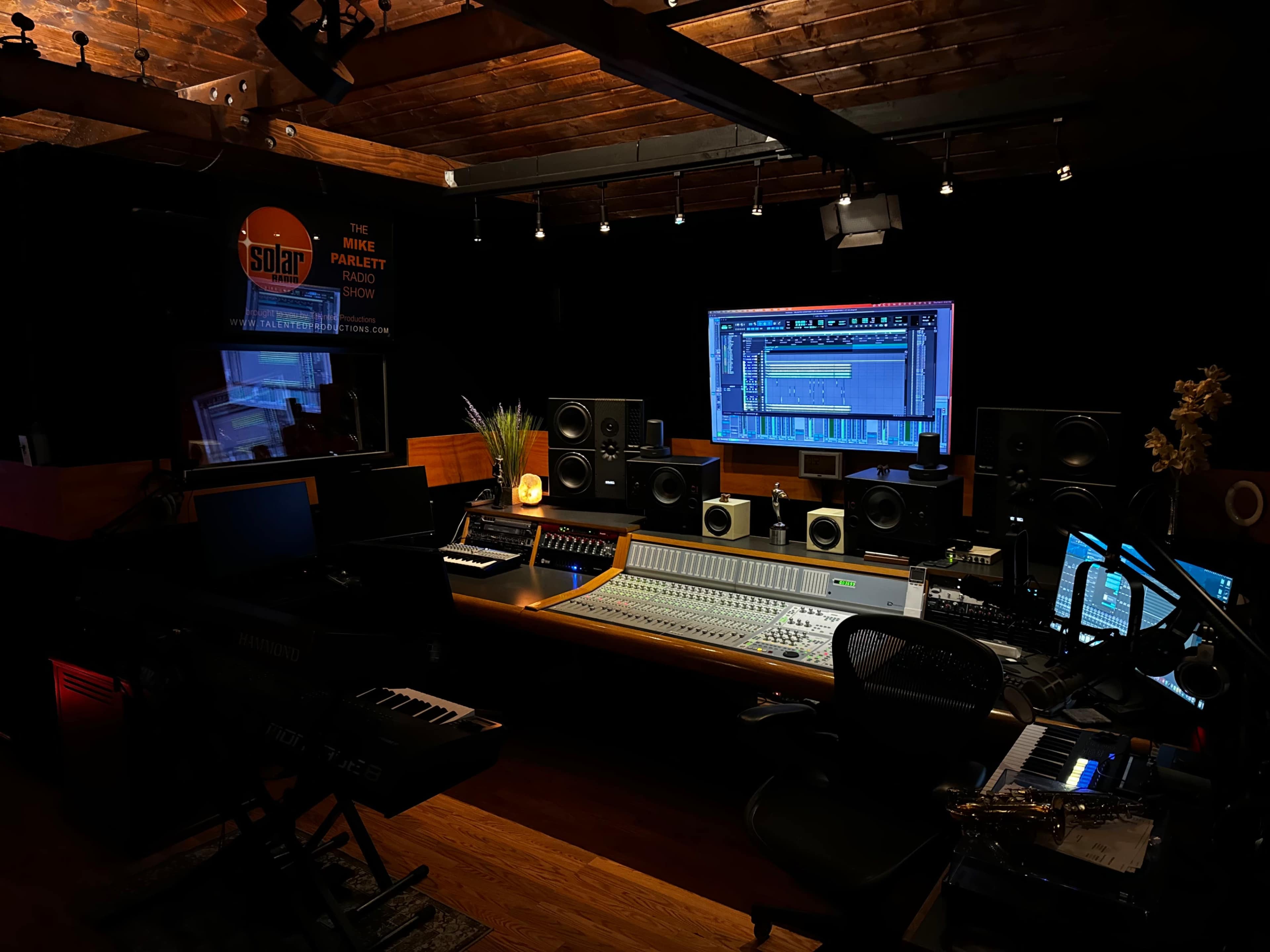 The image shows a music recording studio featuring a large mixing console, multiple speakers, and a computer monitor displaying audio software, all set in a darkened room with wooden beams and ambient lighting.