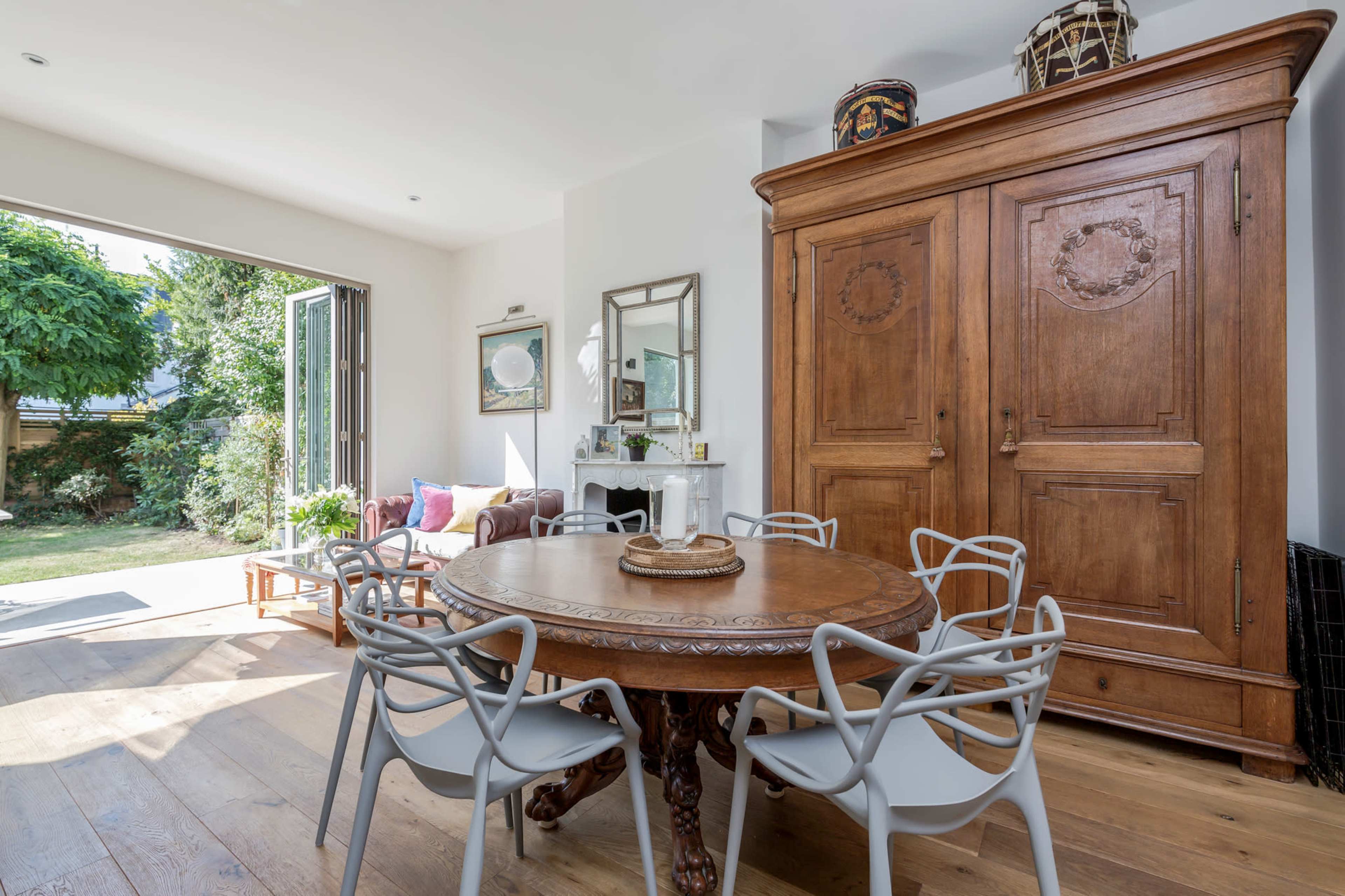 A wooden table surrounded by six chairs is set in a light-filled dining area, with an antique cabinet and large glass doors opening to a garden.