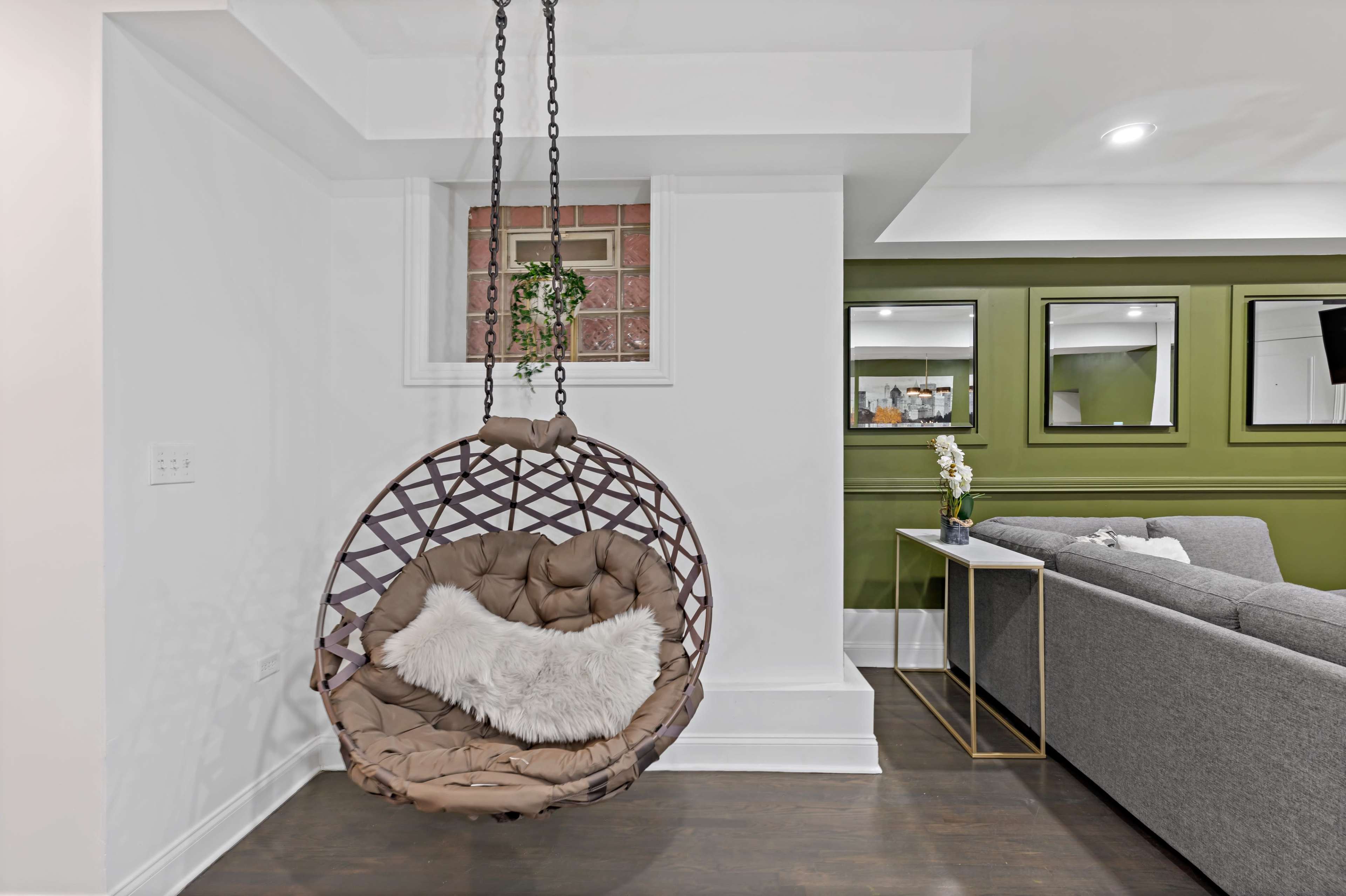 The image shows a cozy interior space featuring a hanging chair with cushions, a small side table, and a gray sofa against a green accent wall.