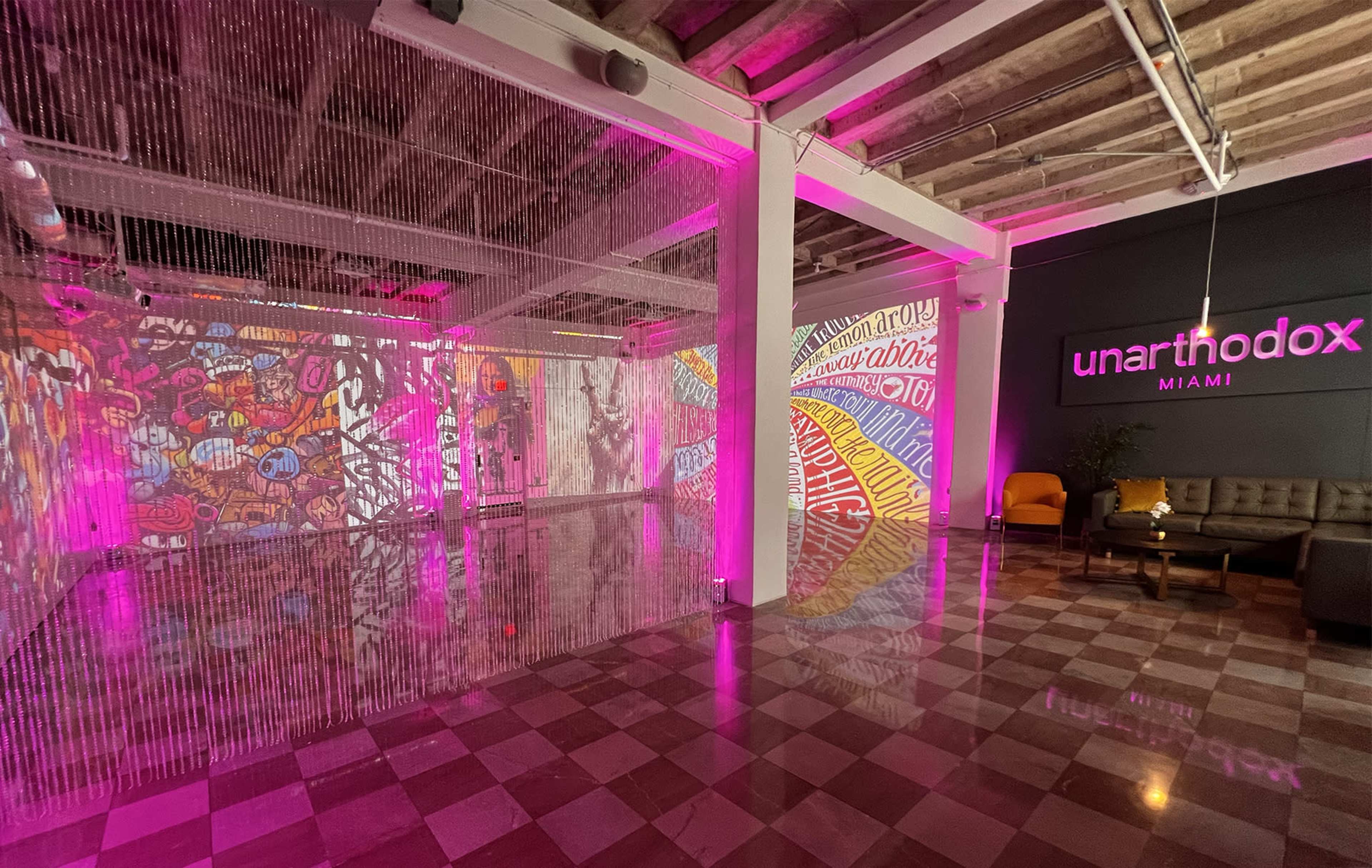 The image shows a brightly lit art space in Miami, featuring colorful murals on the walls and projected designs, with a cozy seating area.
