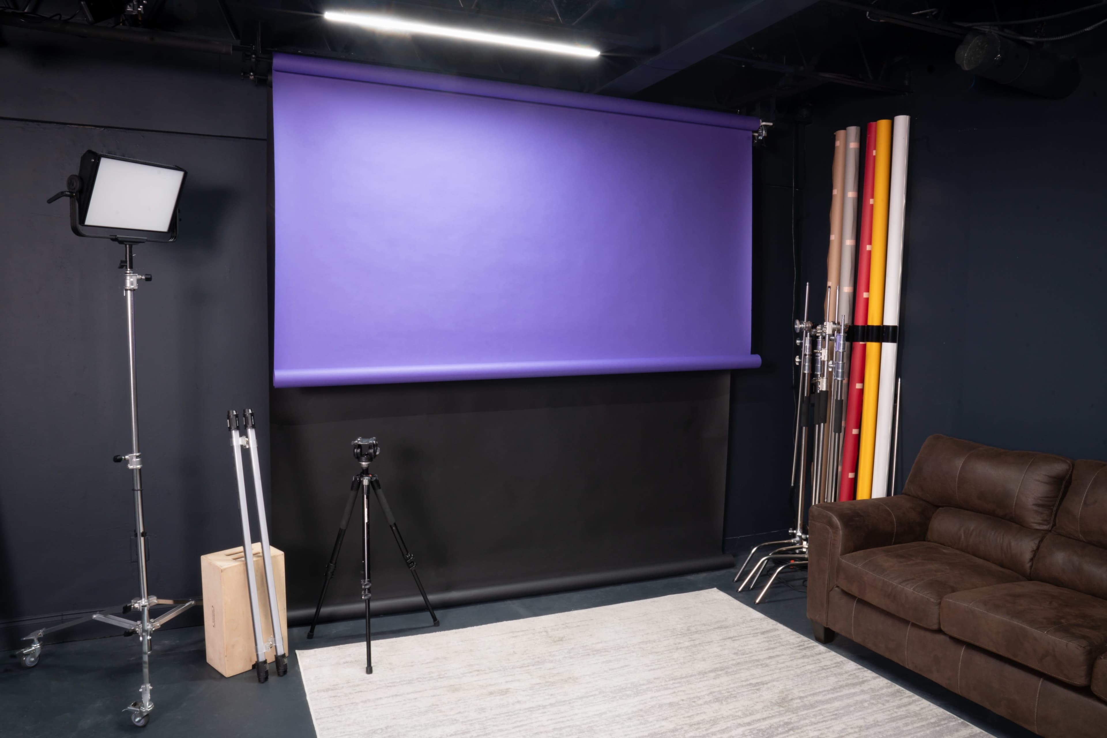The image shows a photography studio with a purple backdrop, a tripod, studio lights, and a brown couch.