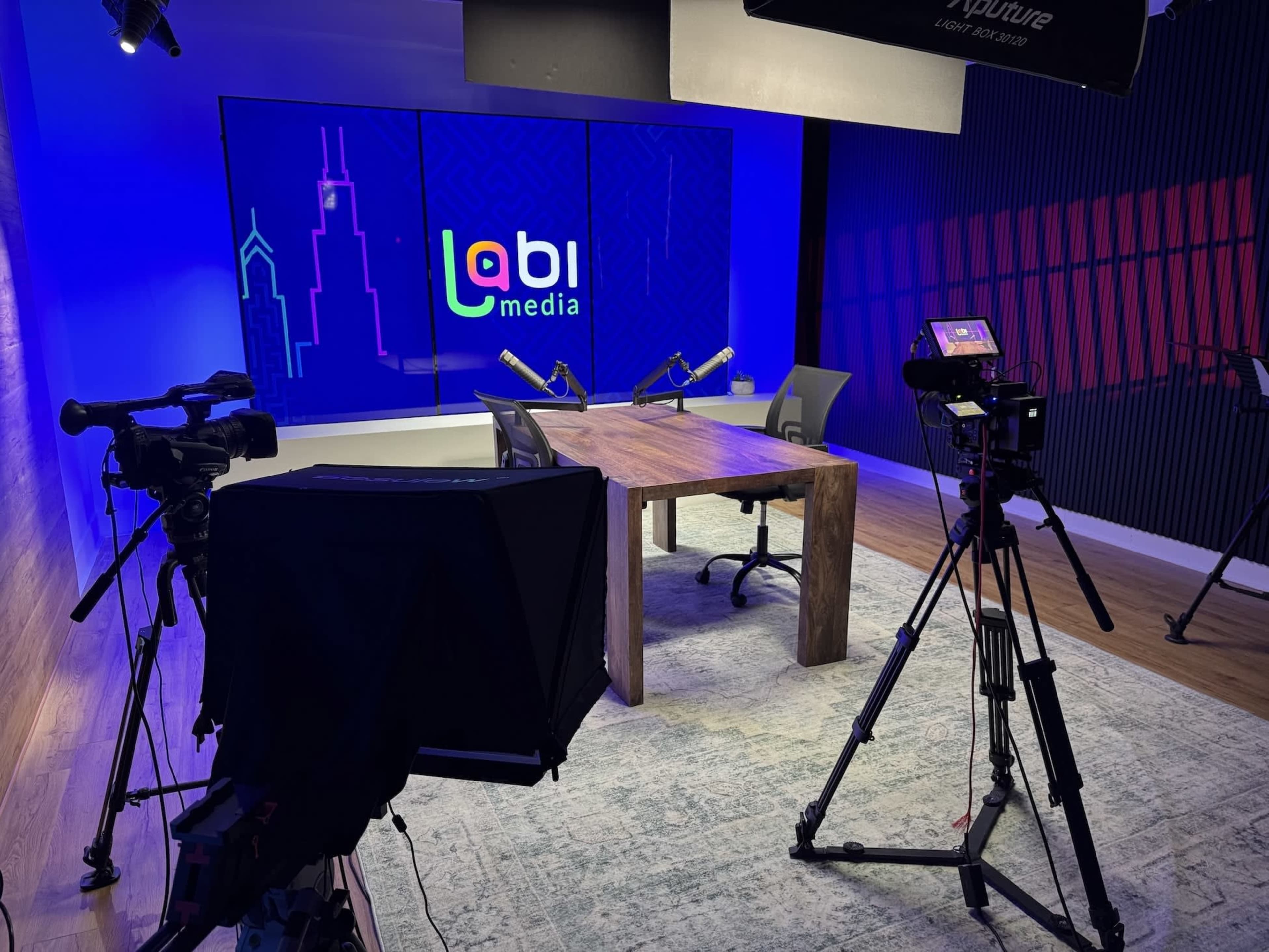 The image shows a professional video production studio with a wooden table, two microphones, and multiple cameras set up for recording, against a backdrop displaying the "Labi media" logo.