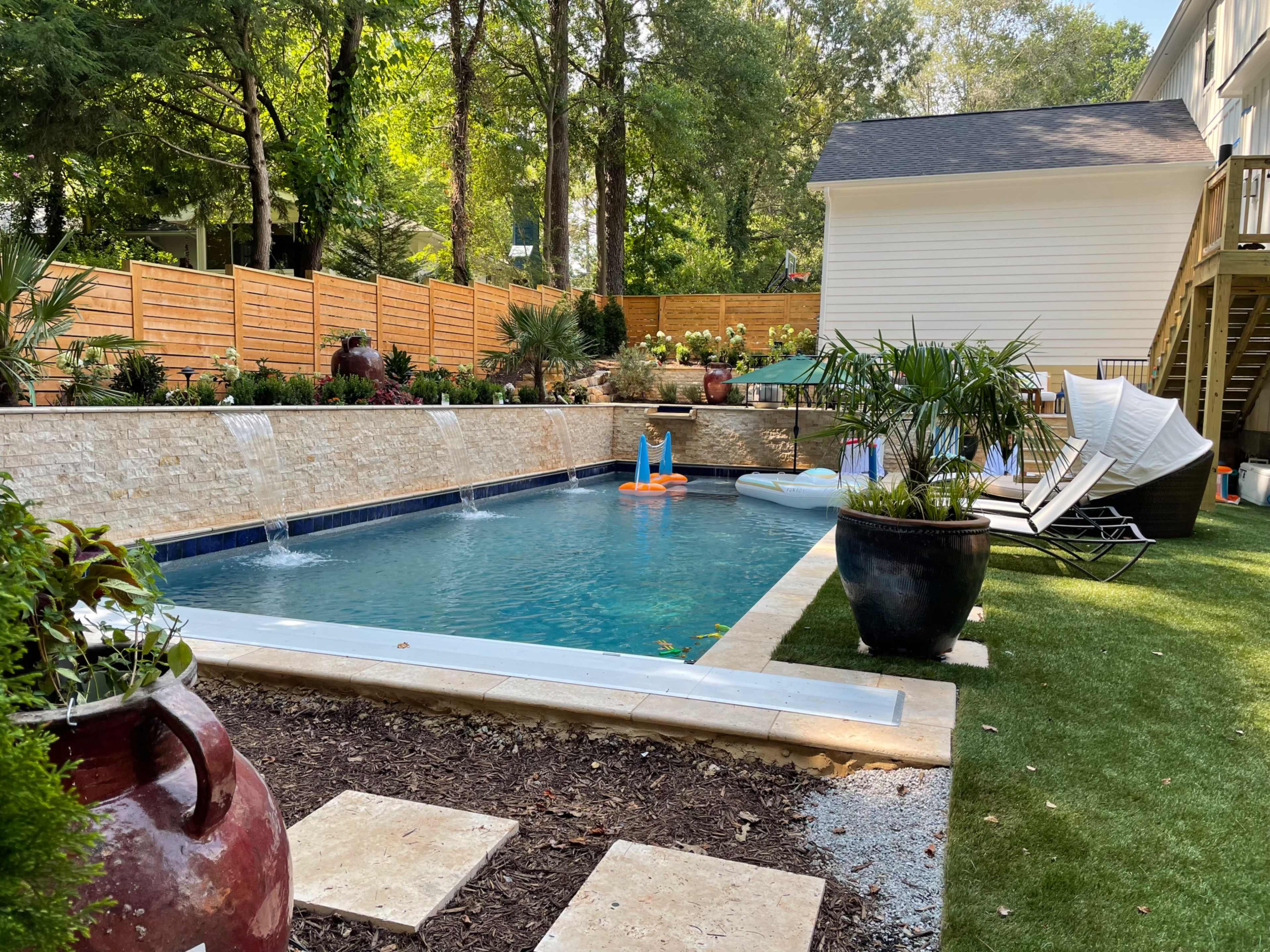 The image shows a backyard pool surrounded by landscaped greenery, deck chairs, and a stone wall with water features, adjacent to a house.