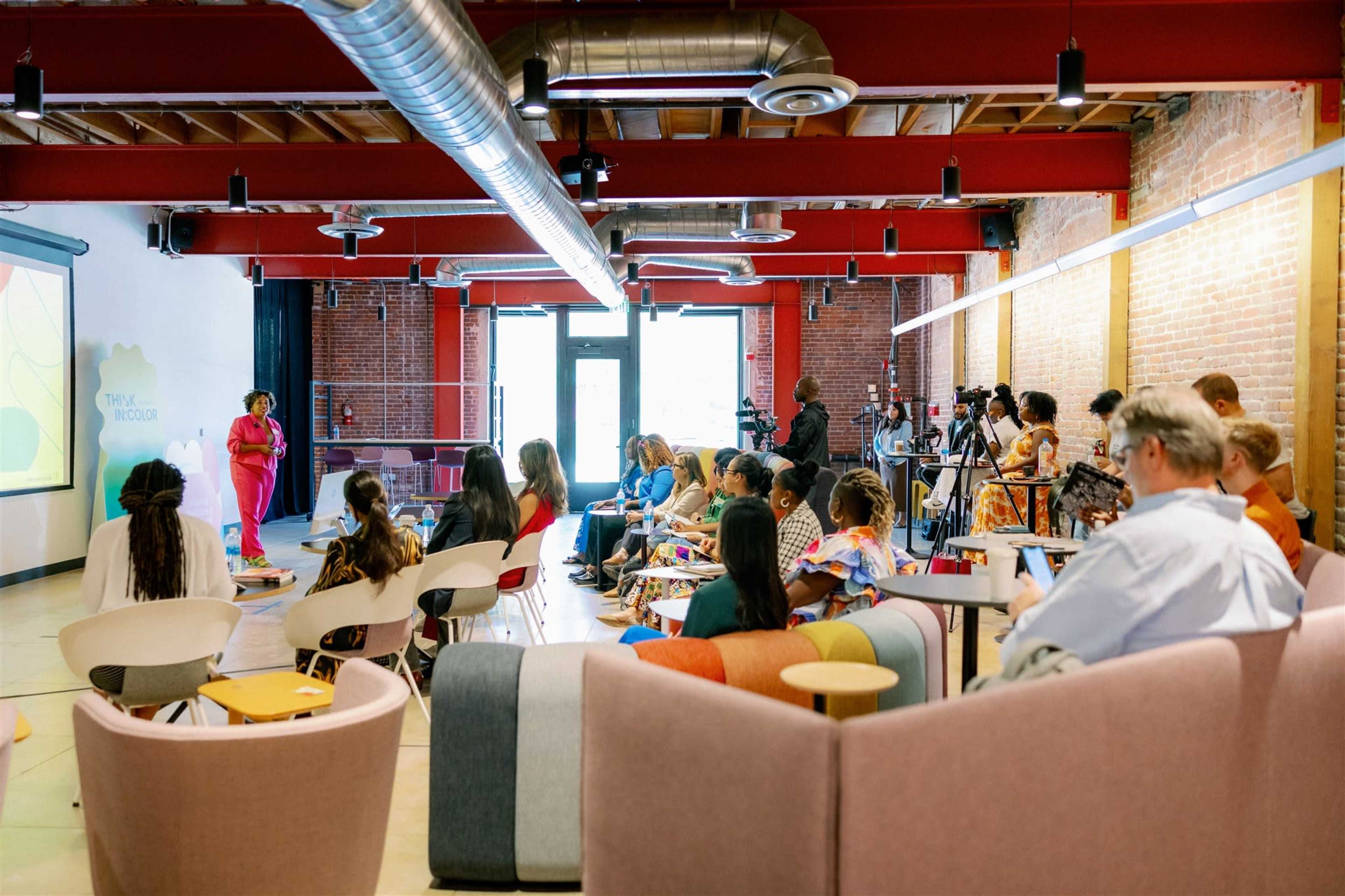 A speaker presents to an audience in a spacious, modern event venue with red beams and exposed brick walls.