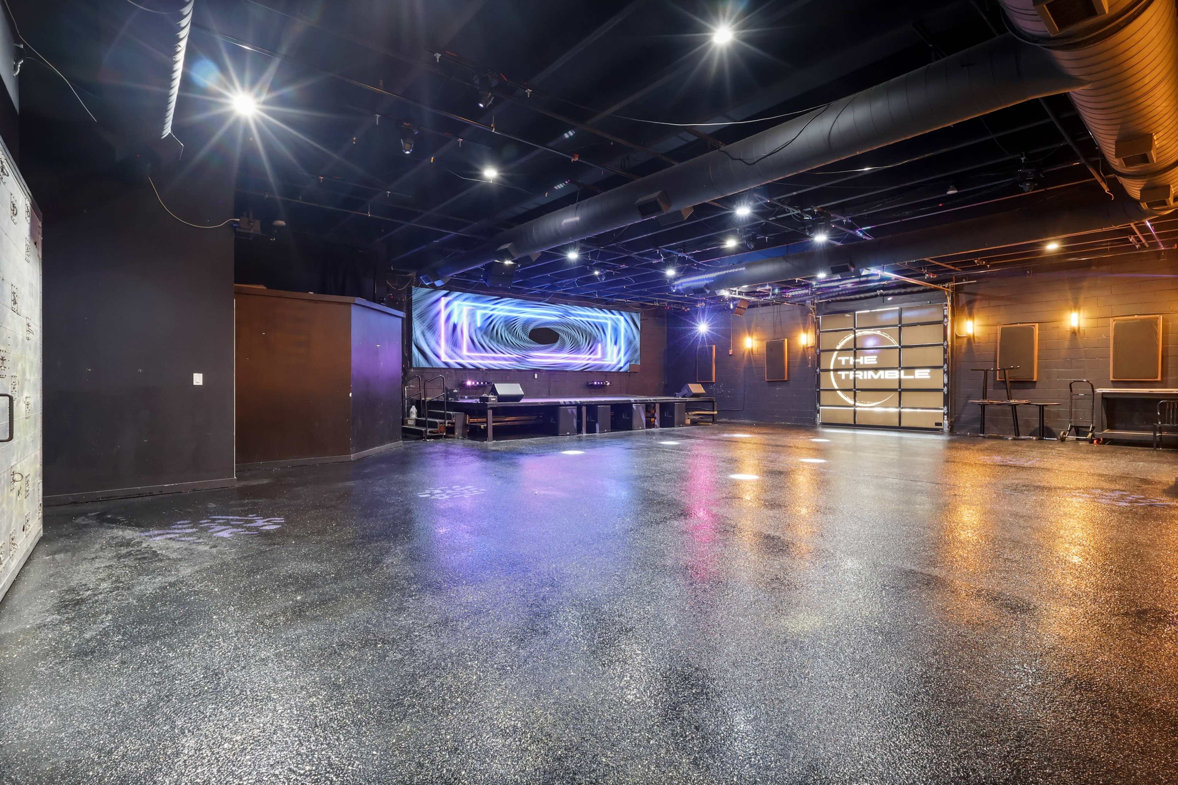 The image shows an empty performance venue with a polished floor, dim lighting, and a backdrop featuring illuminated graphics.