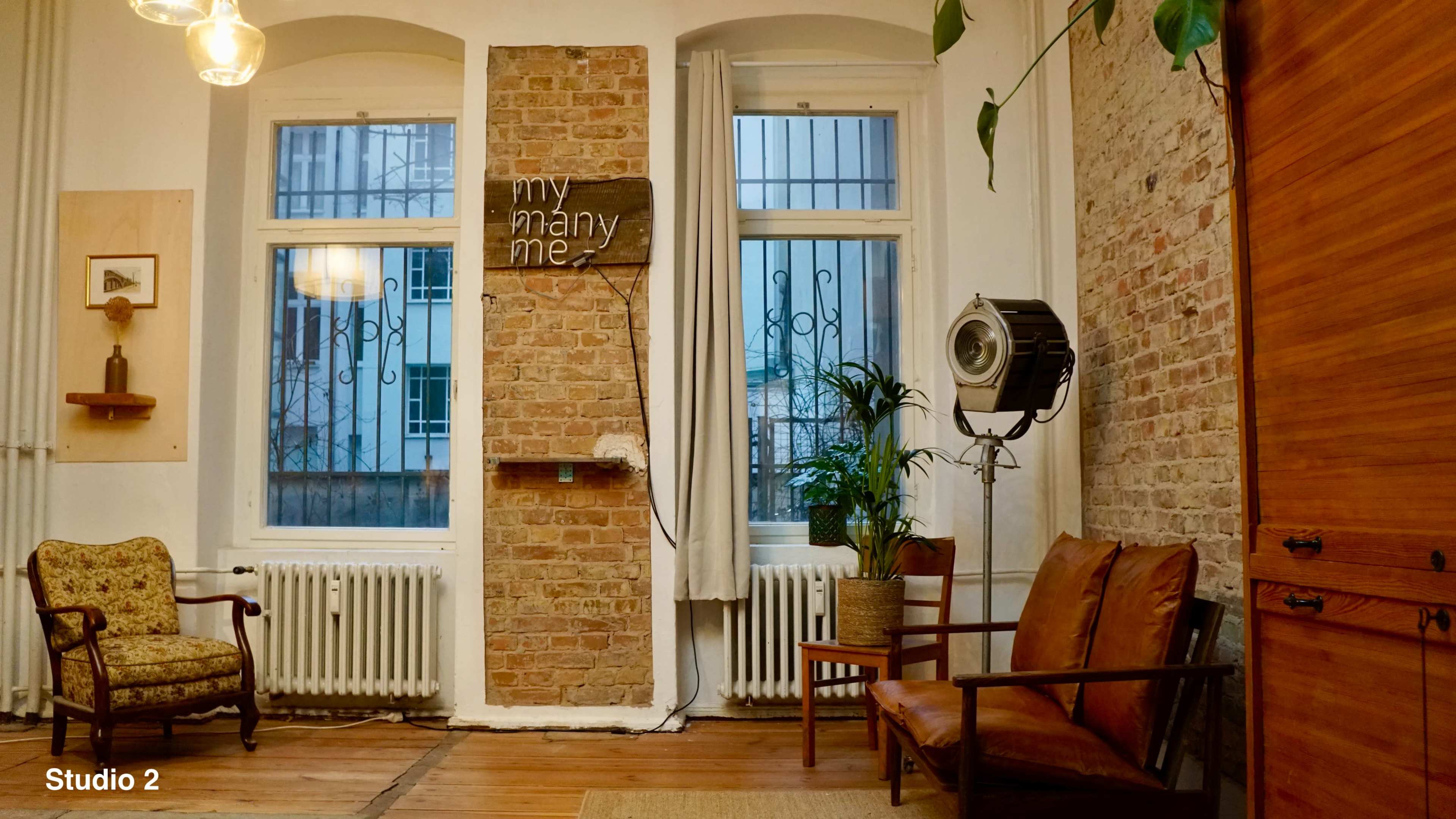 The image depicts a cozy studio space featuring brick walls, a wooden floor, two armchairs, and decorative elements like a plant and a wall sign.