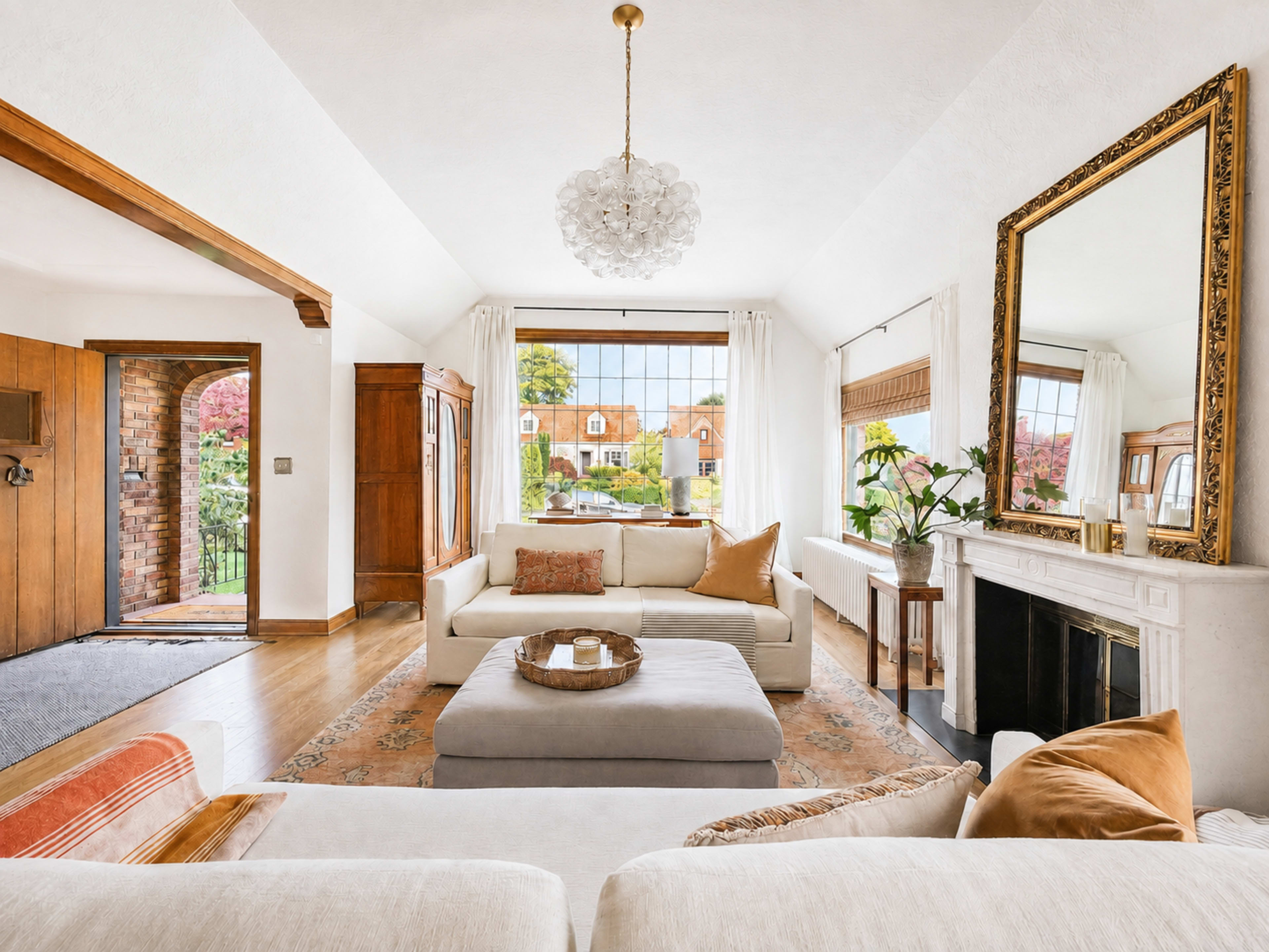 A bright living room features a white sofa with orange cushions, a large mirror above a fireplace, and large windows showing a garden view.