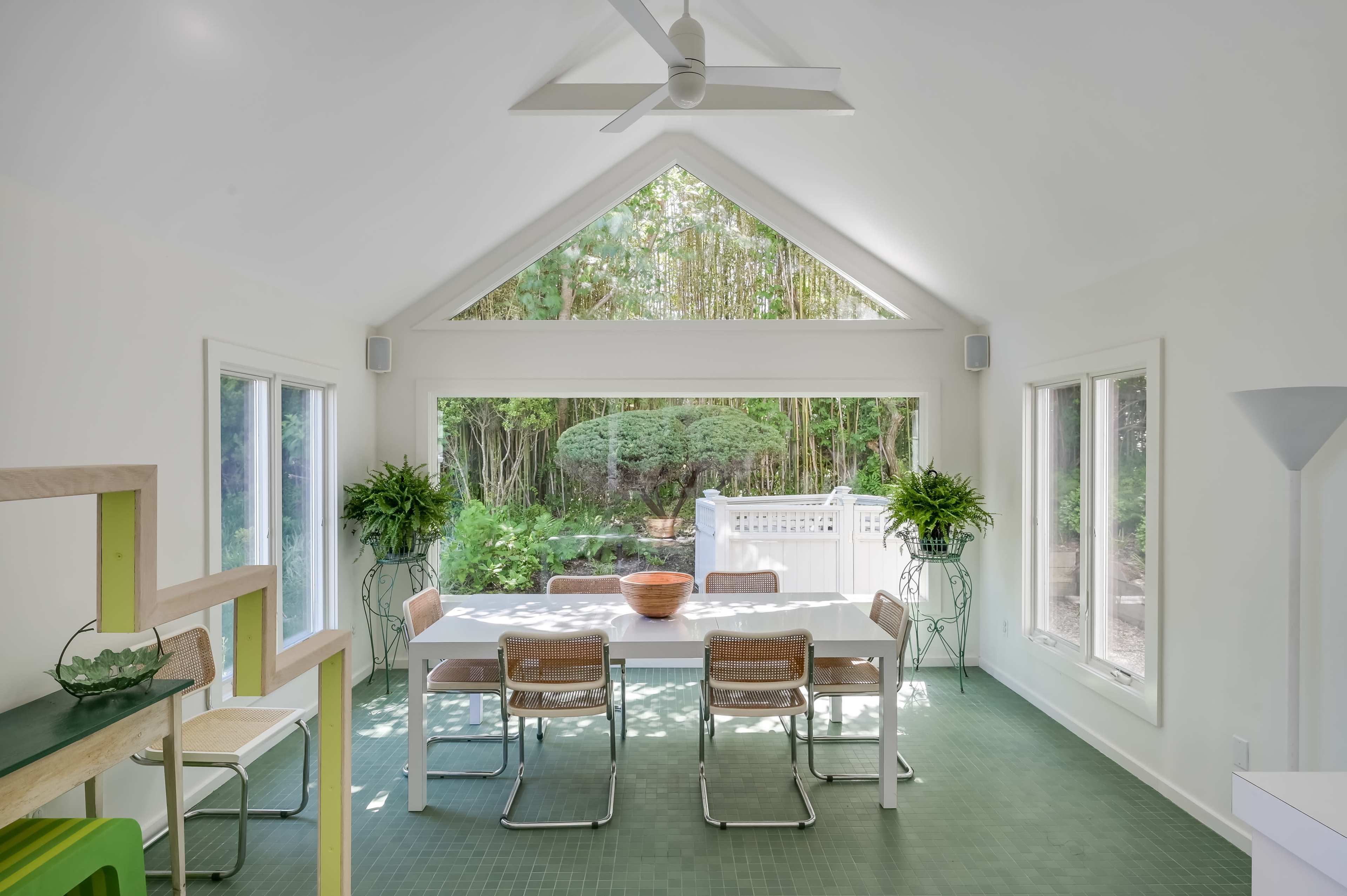A bright dining area with a long table and chairs, surrounded by large windows that overlook a garden.