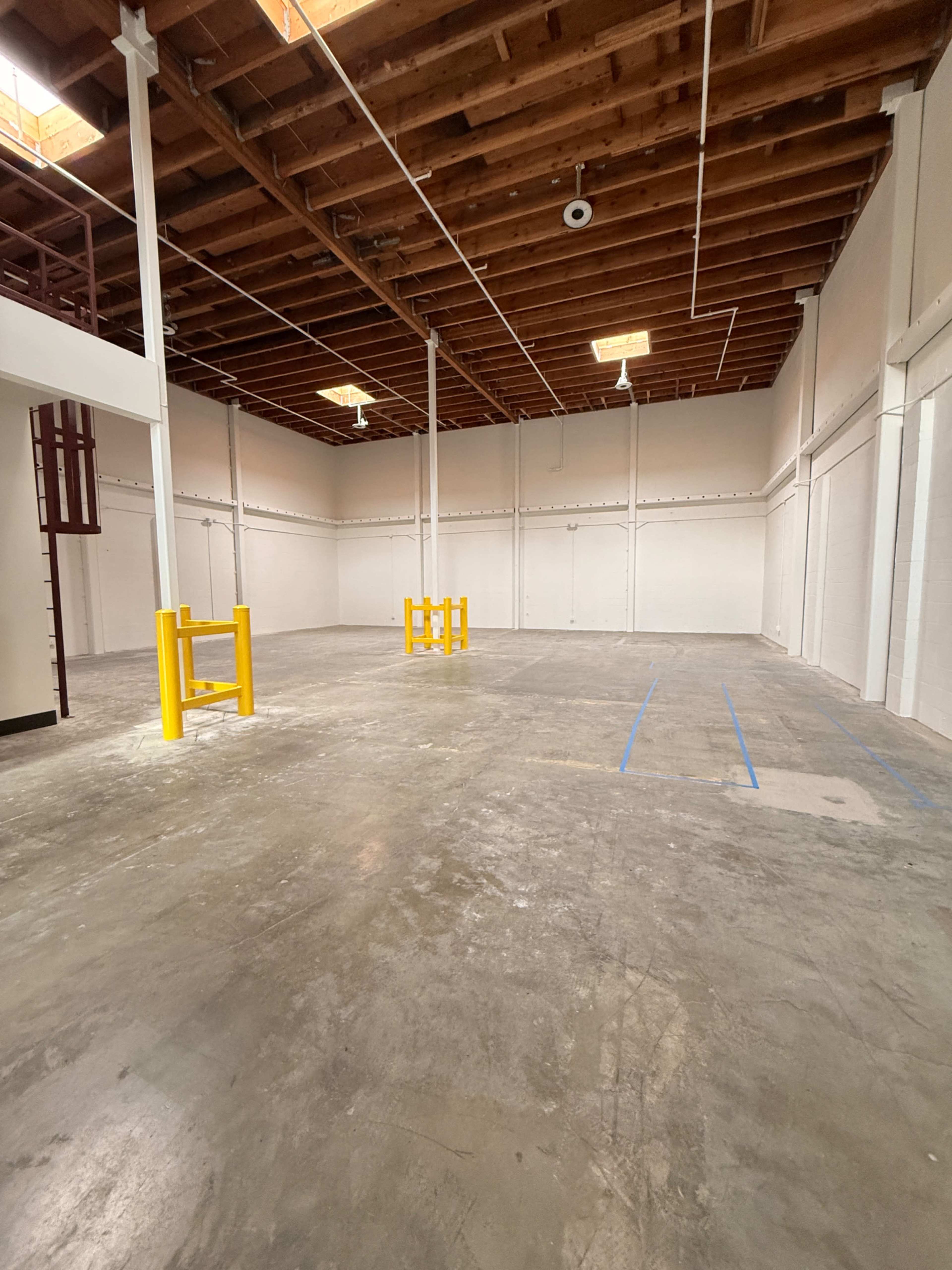 The image shows an empty industrial space with a concrete floor, exposed wooden beams, and yellow safety barriers in the center.