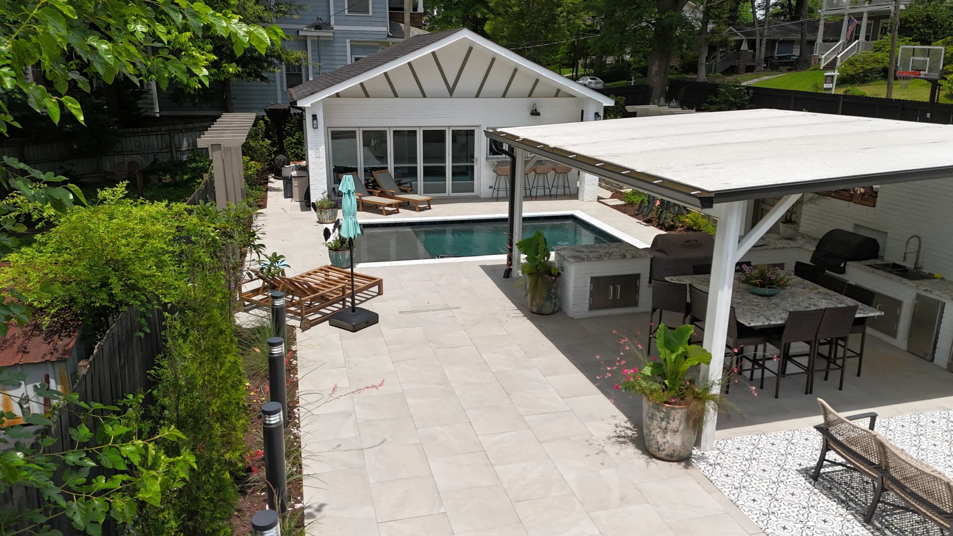 The image shows a backyard featuring a swimming pool, a covered outdoor kitchen area, and a seating arrangement beside a house.