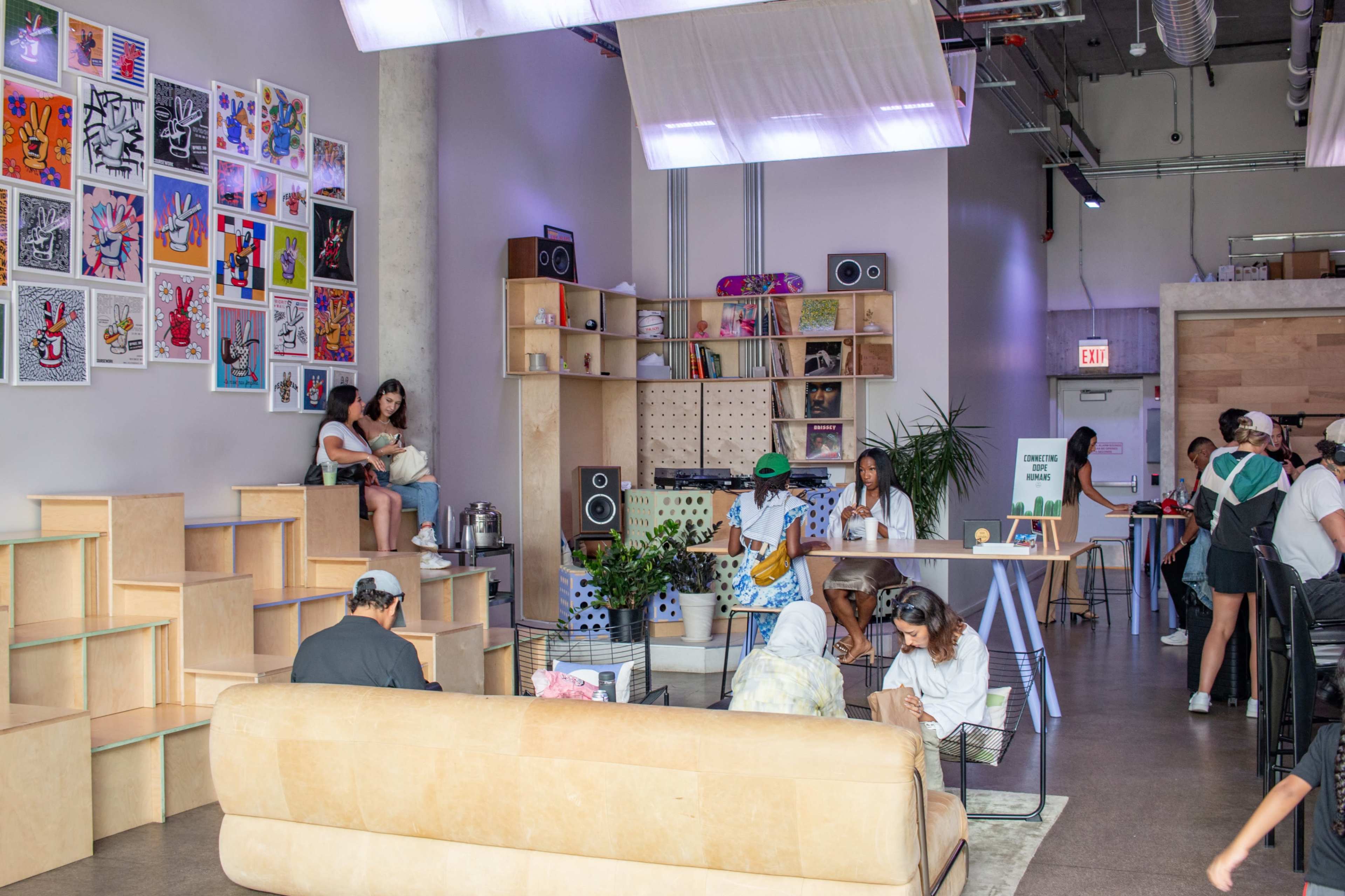 The image shows a modern, open space with varied seating arrangements, colorful artwork on the walls, and people engaging in different activities.