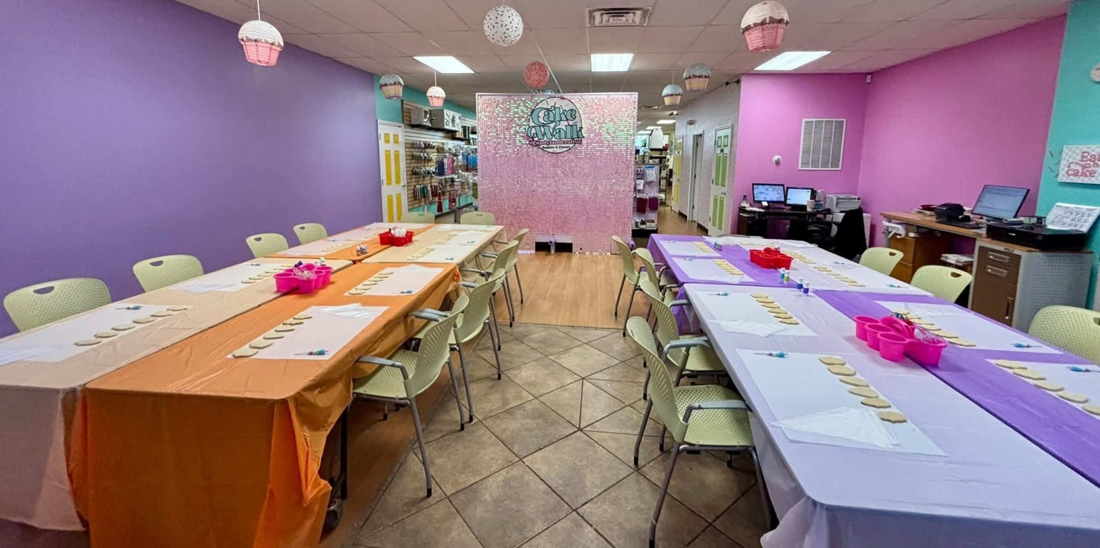 The image shows a colorful craft room with two long tables covered in orange and purple tablecloths, set up for an activity with cookies and supplies on each table, and decorative lanterns hanging from the ceiling.