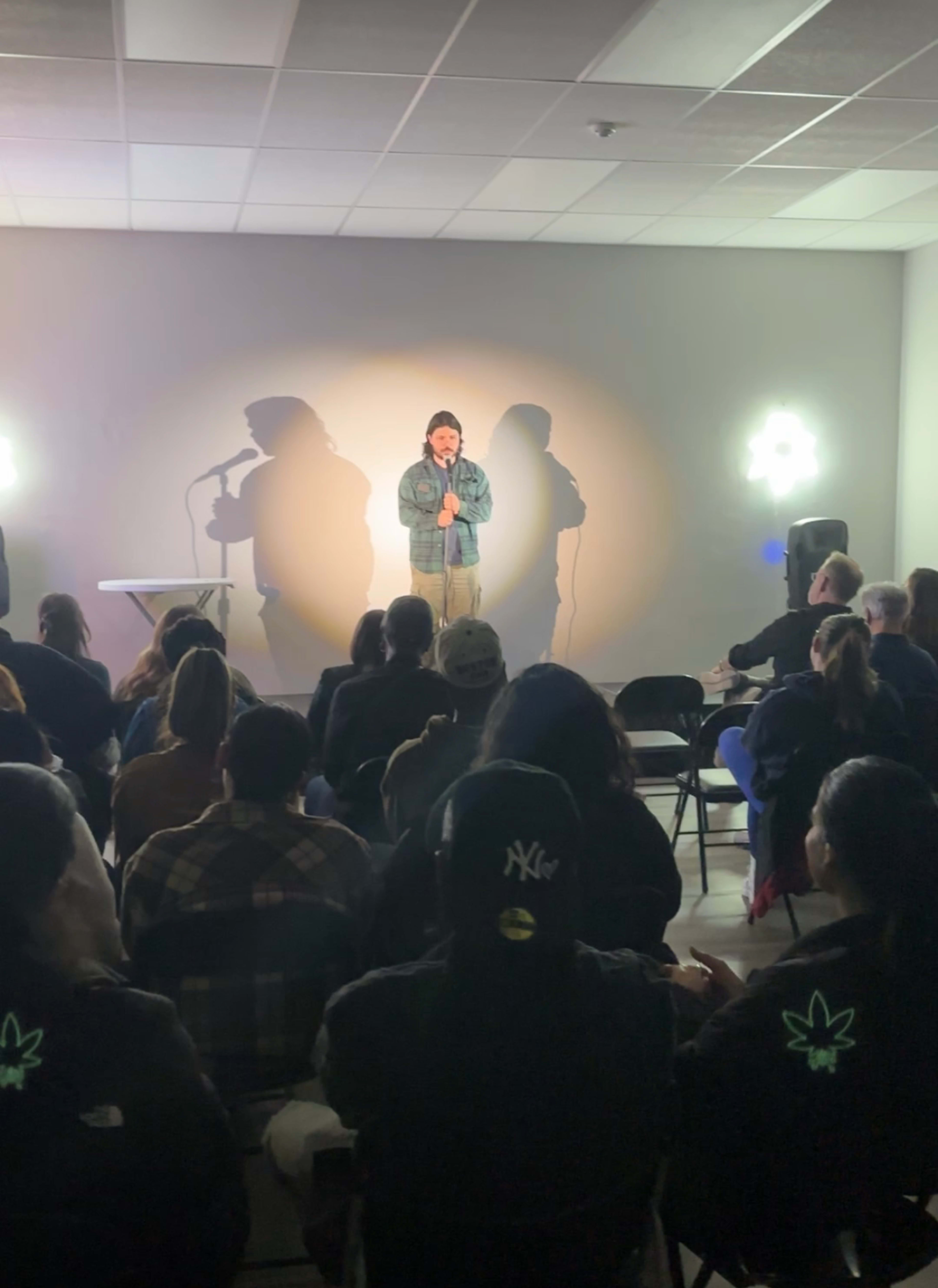 A performer stands on stage in front of an audience seated in chairs, with soft lighting casting shadows on the wall behind them.