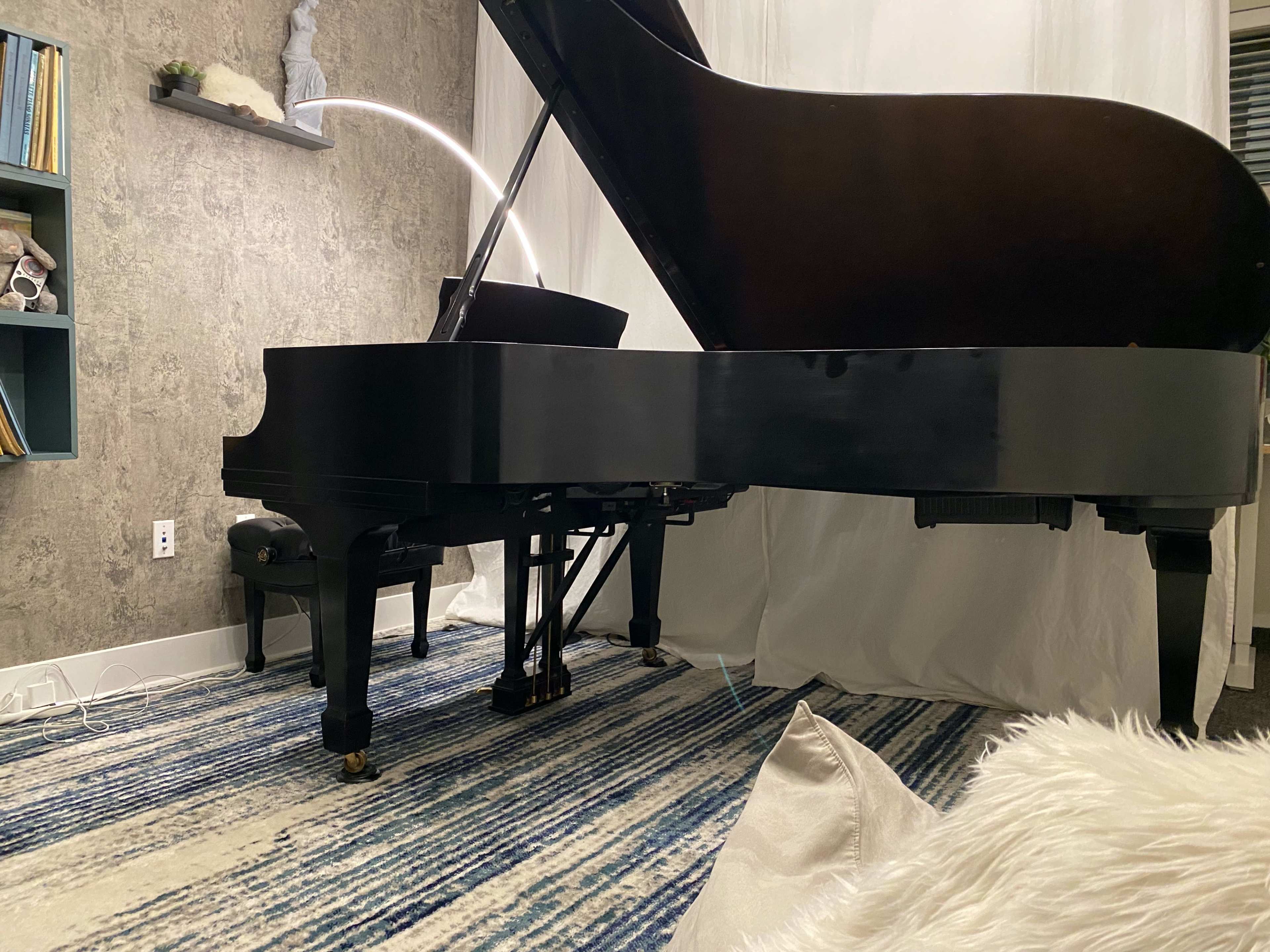 A grand piano is positioned on a patterned rug in a softly lit room with a textured wall and a decorative shelf.