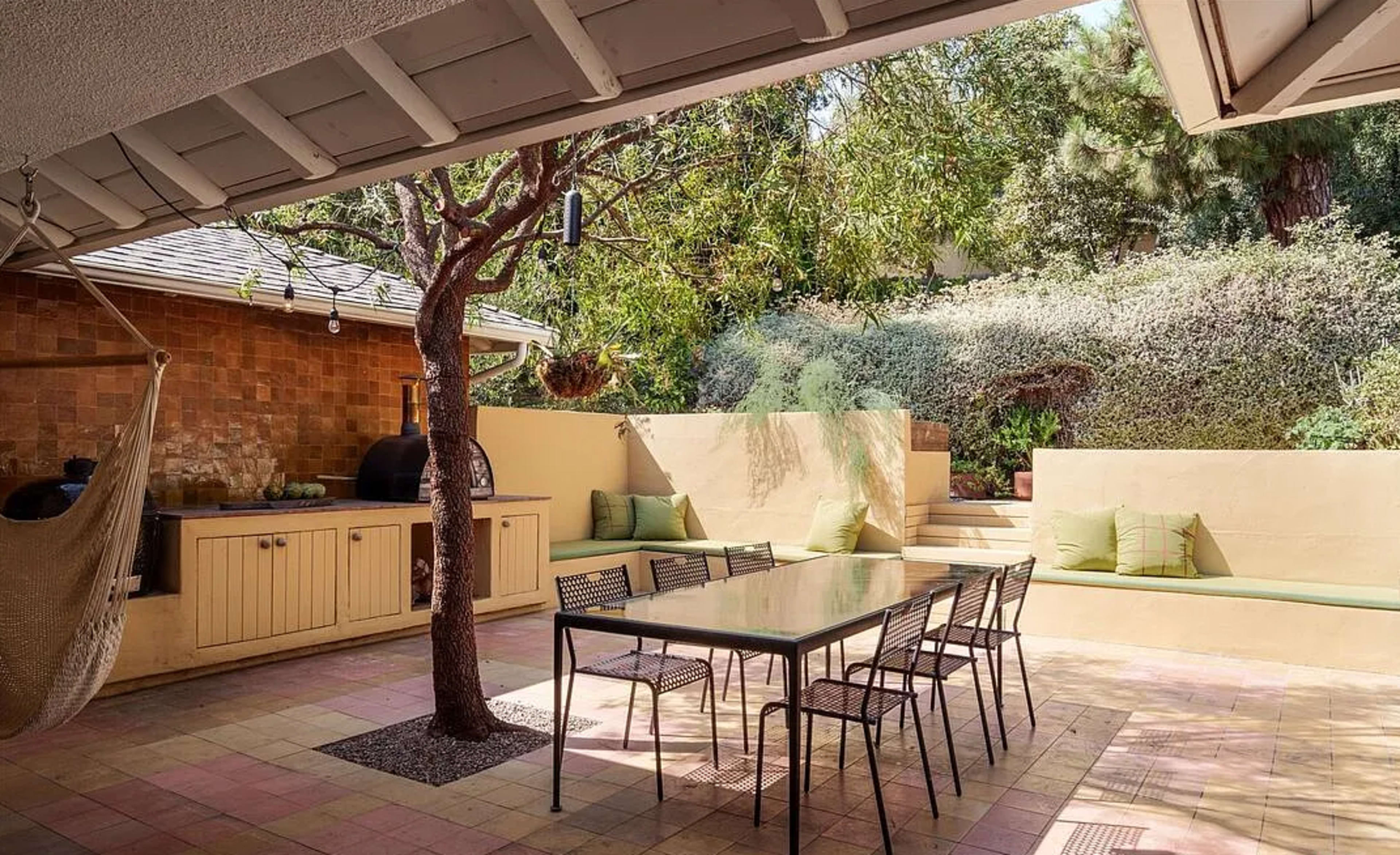 The image shows a landscaped outdoor patio area featuring a dining table with chairs, a tree, a hammock, and seating along the walls.