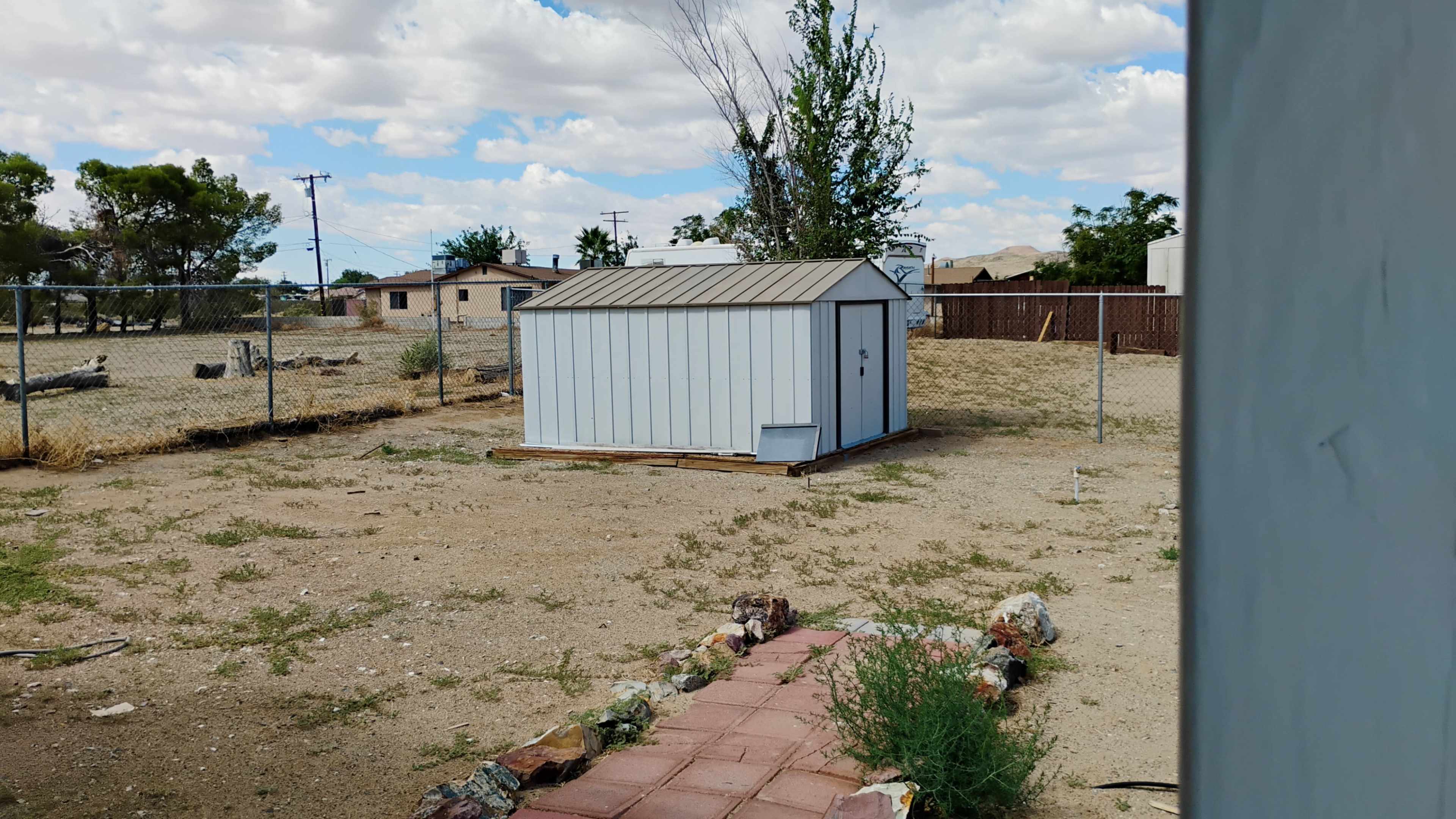 The image shows a small gray storage shed in a barren backyard with dry grass and scattered shrubs, surrounded by a fence and houses in the background.